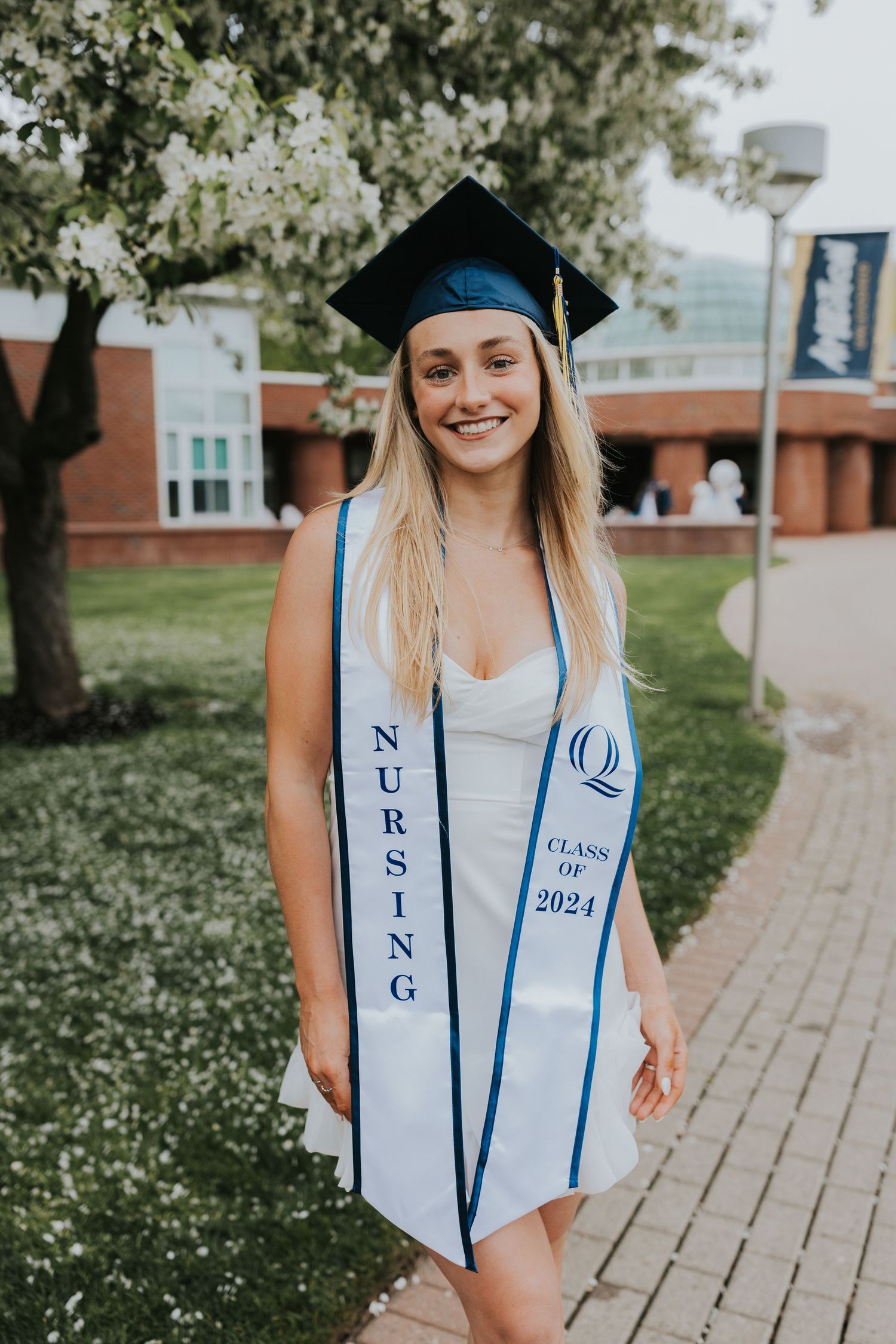 Smiling nursing graduate in cap and white dress with Class of 2024 stole on a brick campus path.