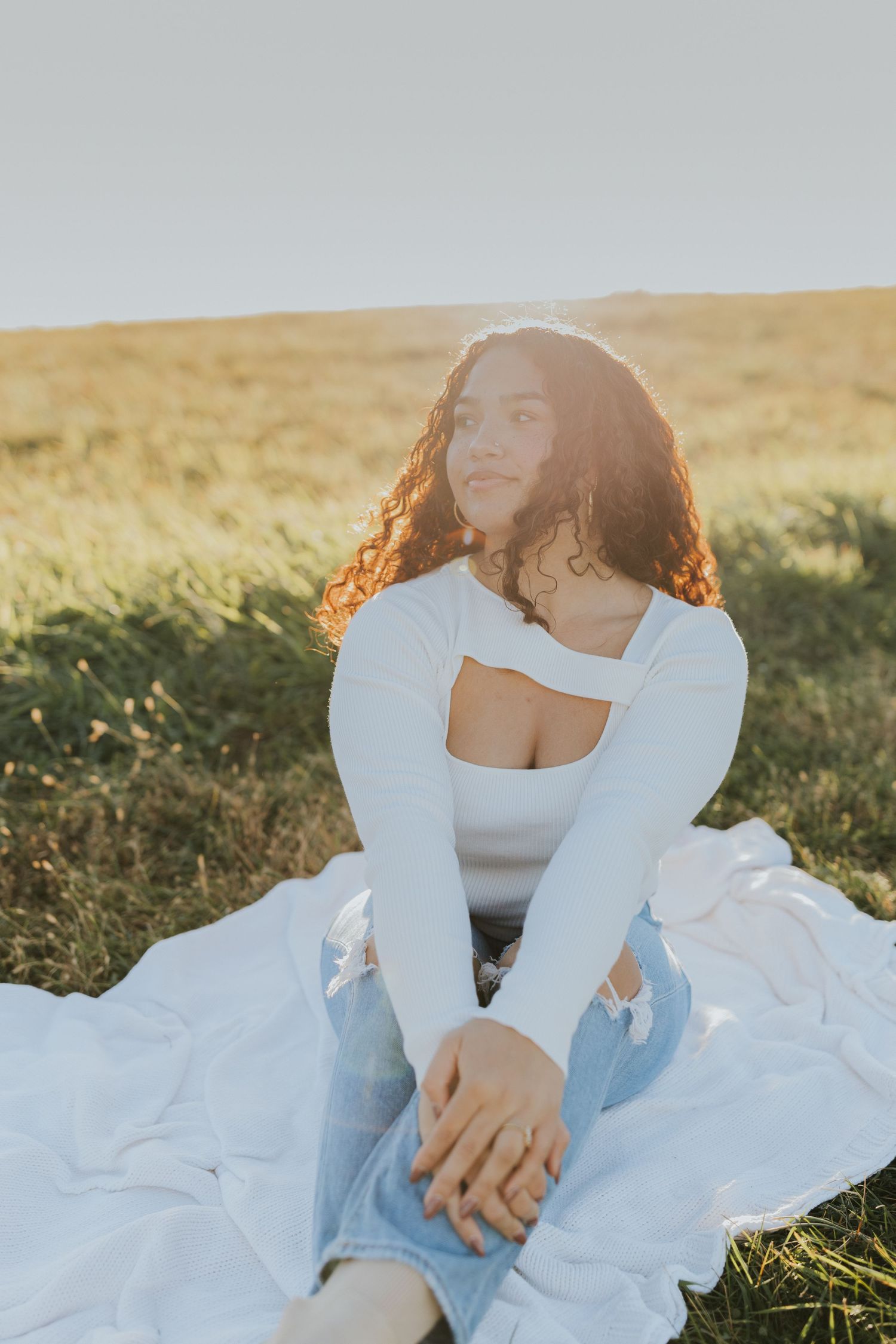 Young woman with curly hair sitting on white blanket in golden hour sunlit grassy field.