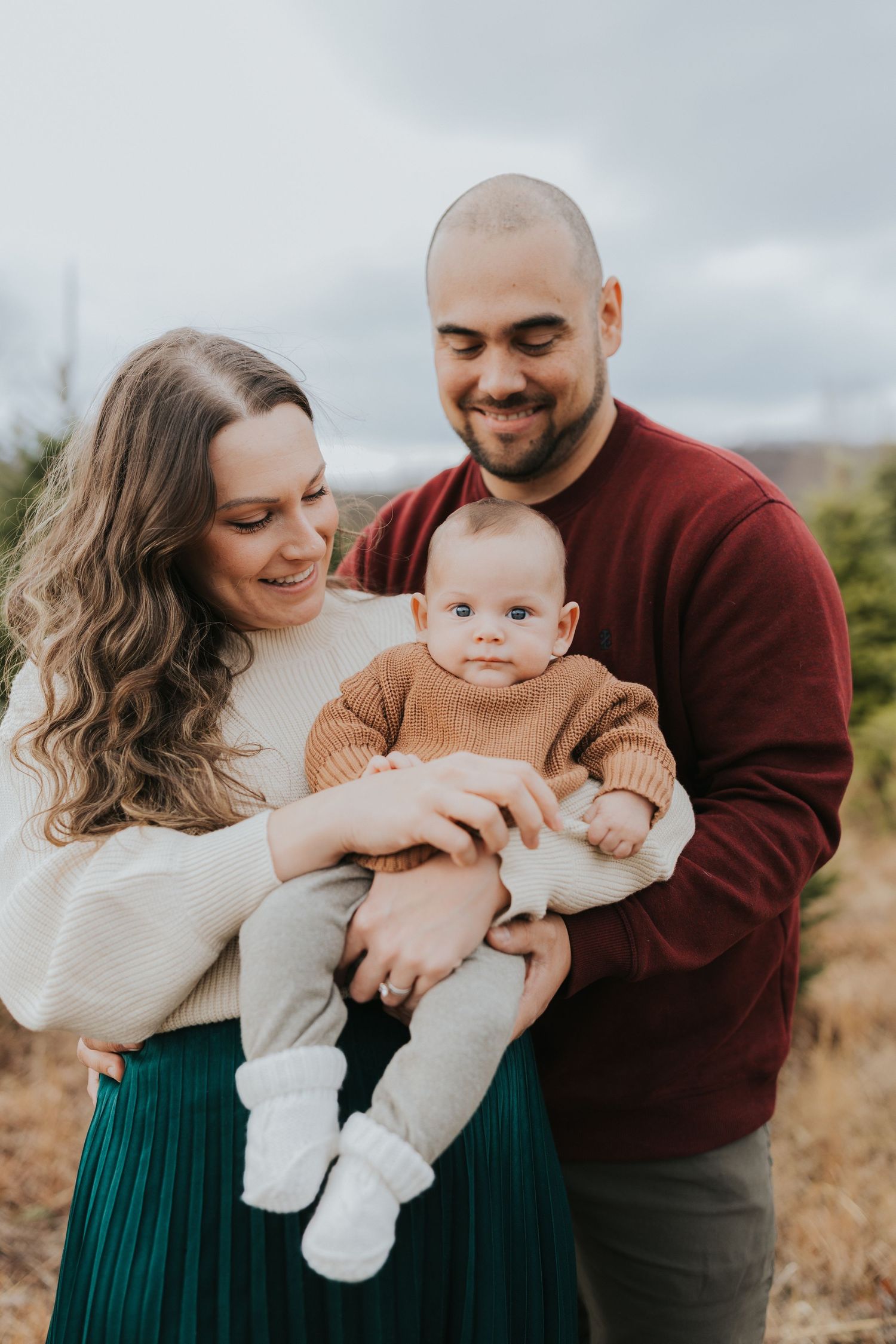 Parents in cream and burgundy sweaters smile down at baby held between them at tree farm.