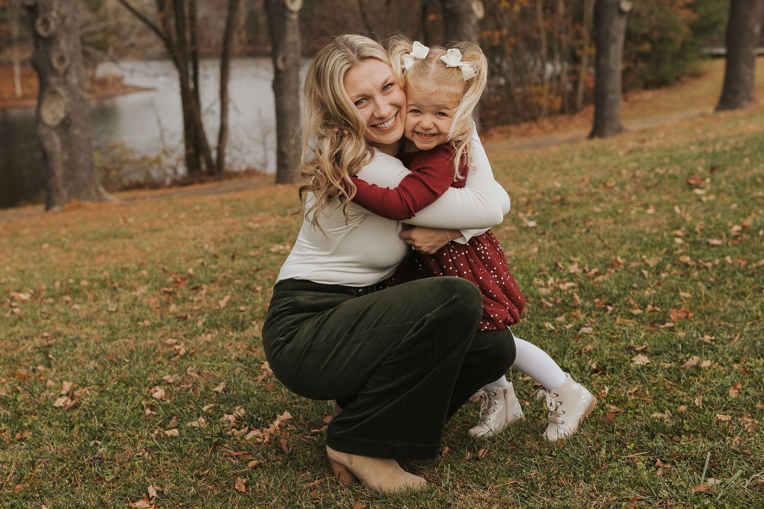Blonde mother crouching down to hug her smiling toddler daughter outdoors in fall foliage.