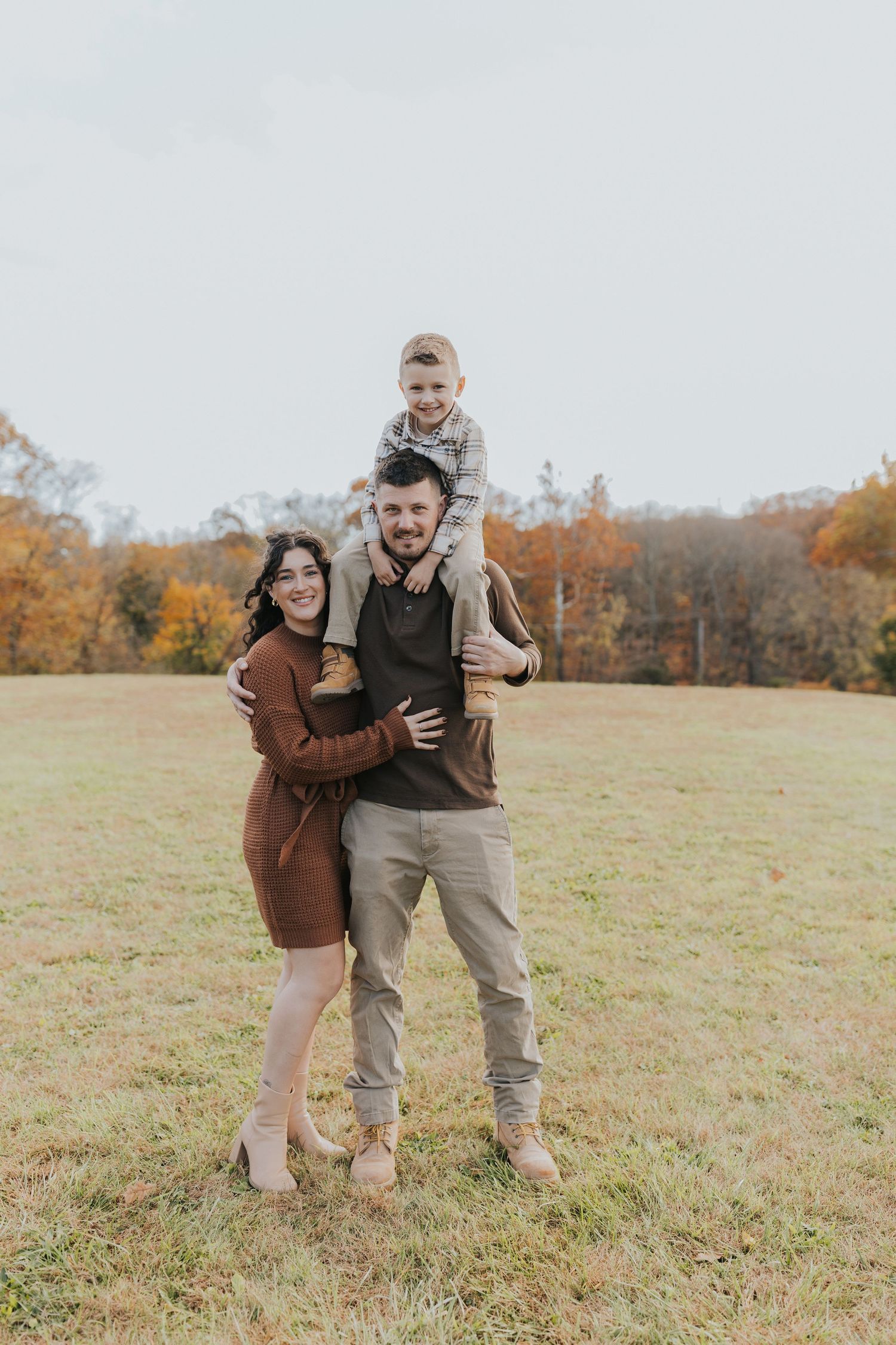 A family of three poses in an open field with autumn foliage, son perched on father's shoulders.