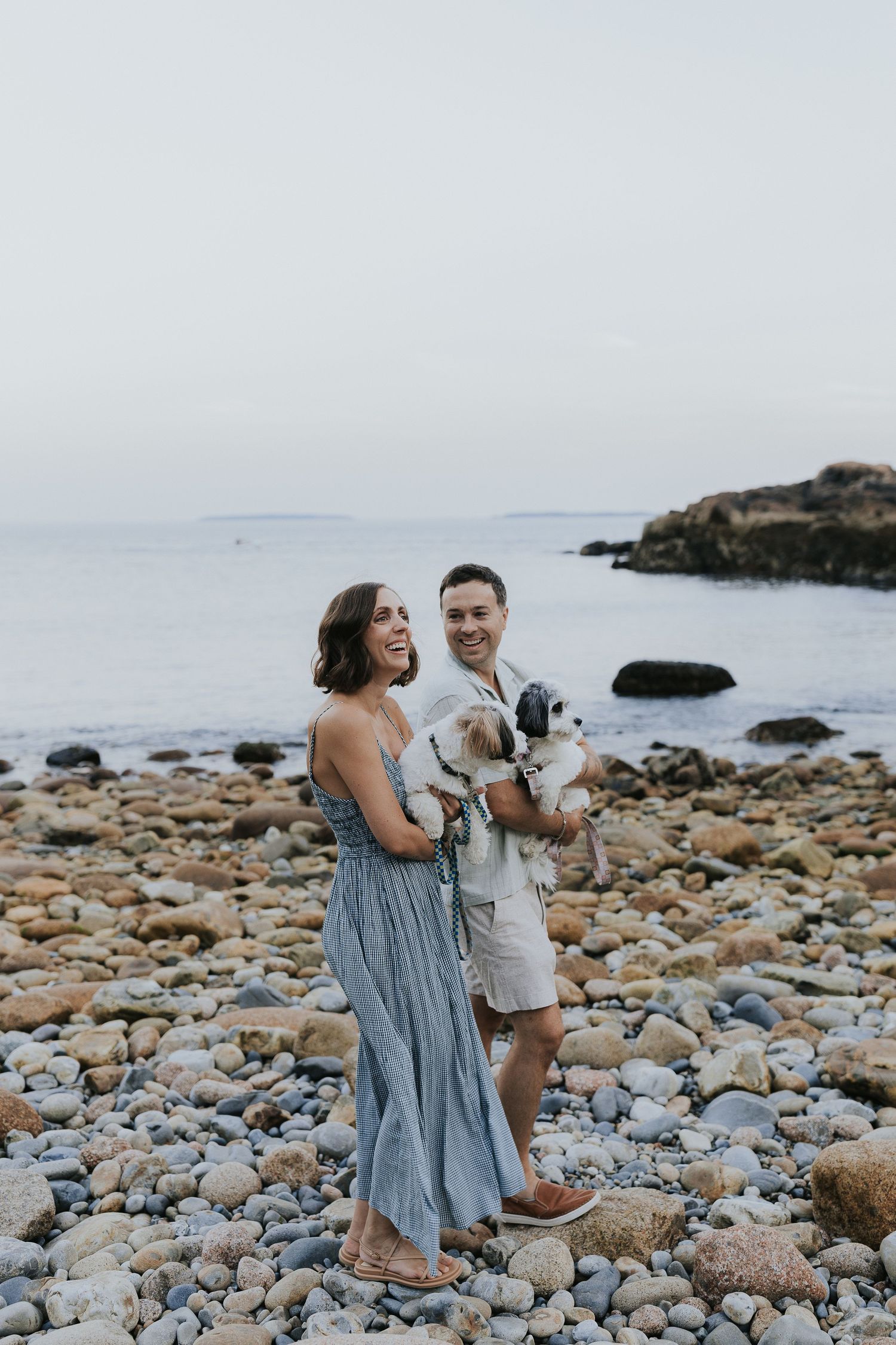 Happy couple holding a small dog on a pebble beach with ocean views.