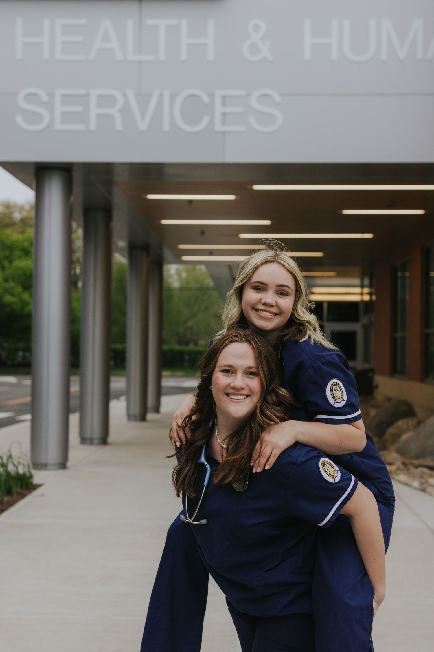 Two nursing students in navy scrubs pose playfully outside a Health and Human Services building.