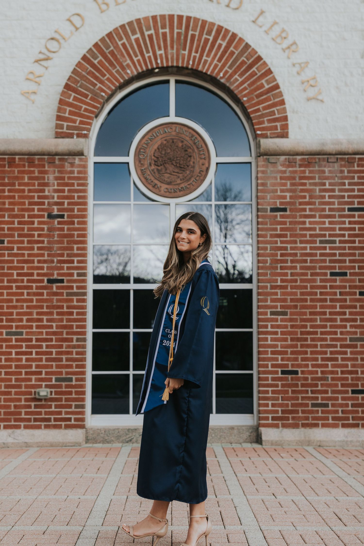 Graduate in navy gown with nursing stole stands sideways at Arnold Bernhard Library brick entrance.