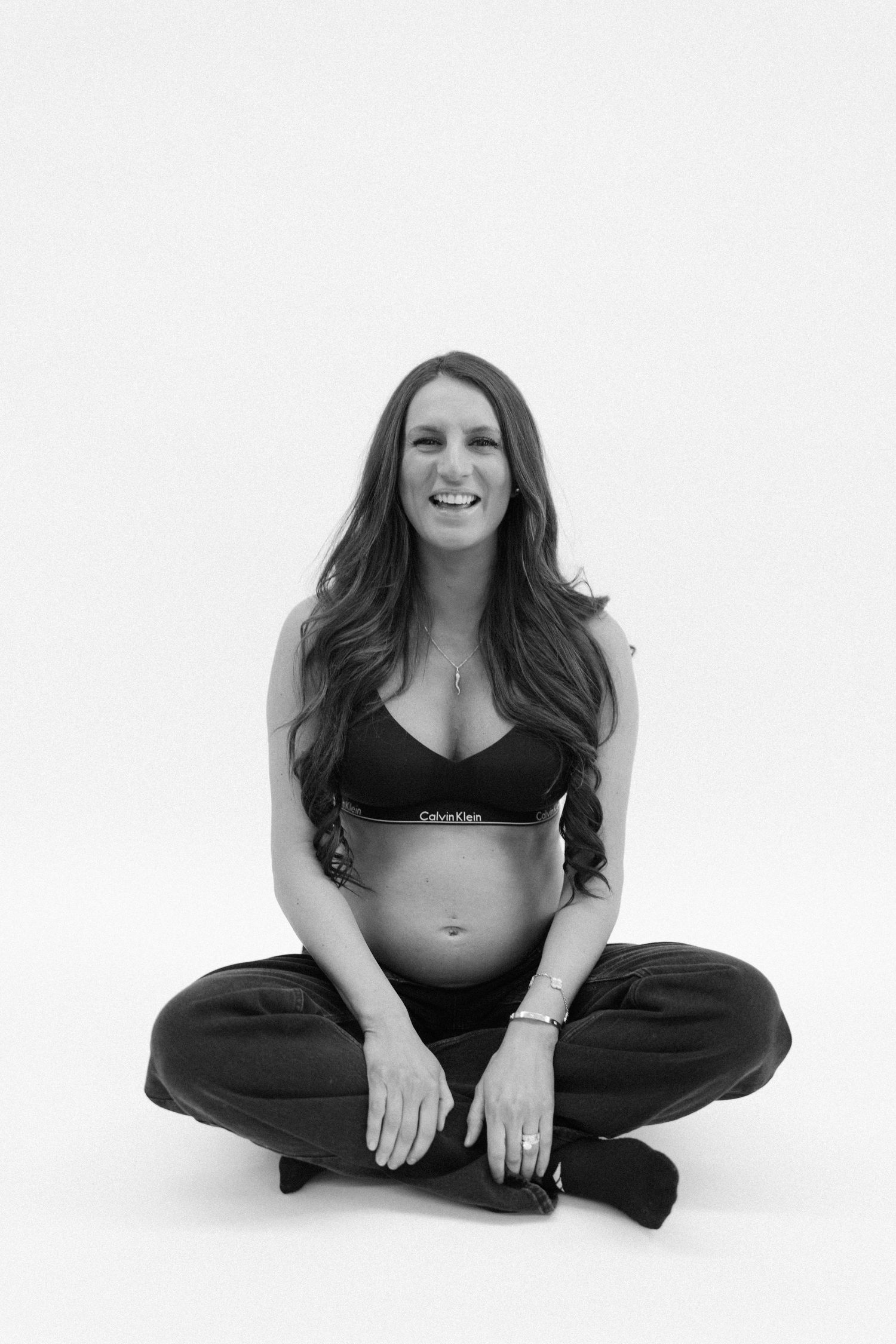 Smiling pregnant woman in Calvin Klein bralette sitting cross-legged in black and white photo.