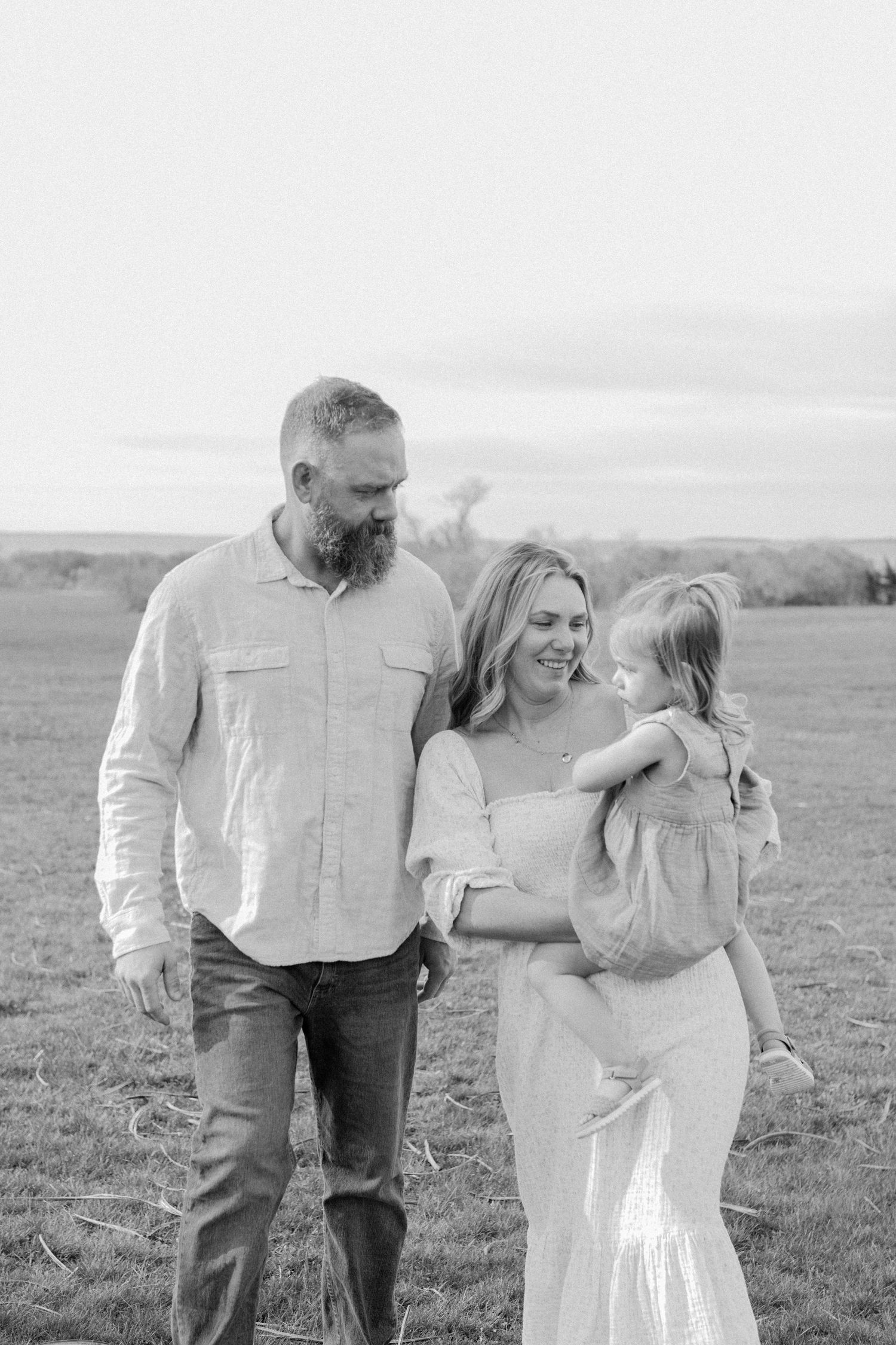 Black and white photo of bearded father, smiling mother holding toddler girl in open field.