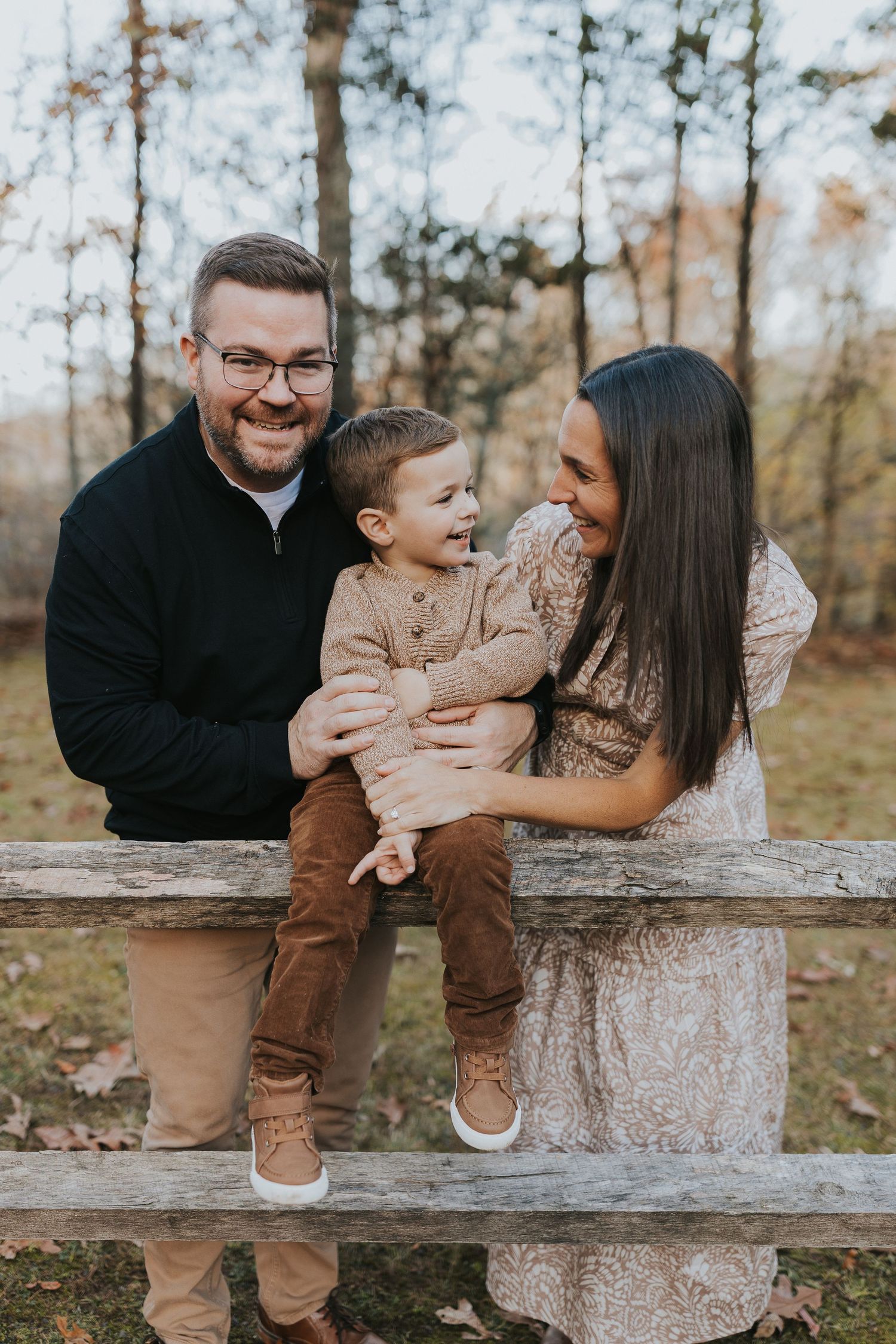 A joyful family of three laughs together while sitting on a rustic wooden fence in autumn woods.