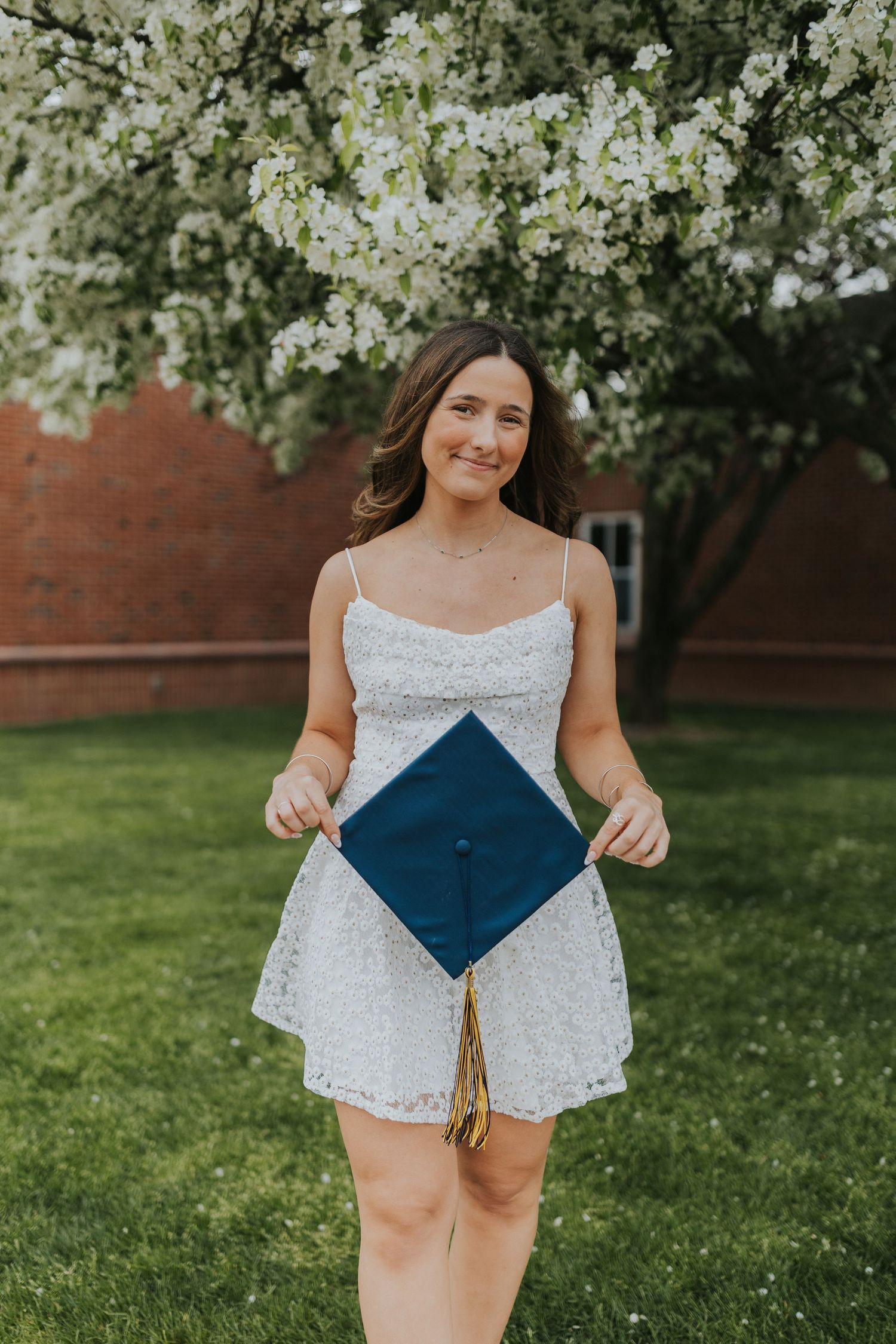 Graduate in white lace dress holds navy graduation cap while standing on green lawn near blooming tree.