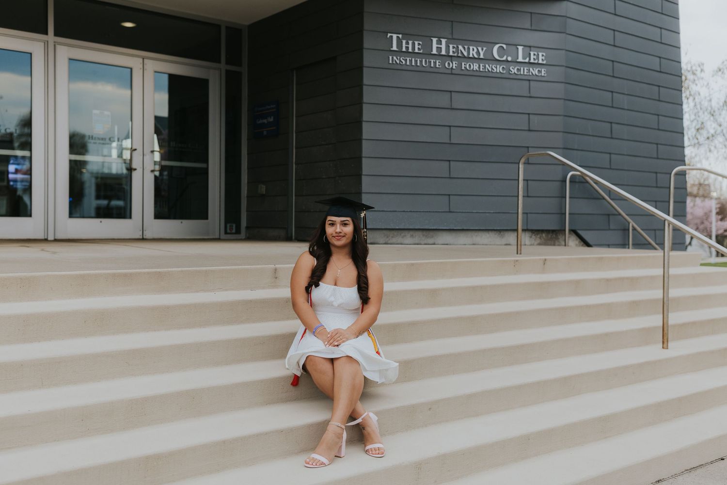 Graduate in white dress and cap sits on steps of Henry C. Lee Institute of Forensic Science.