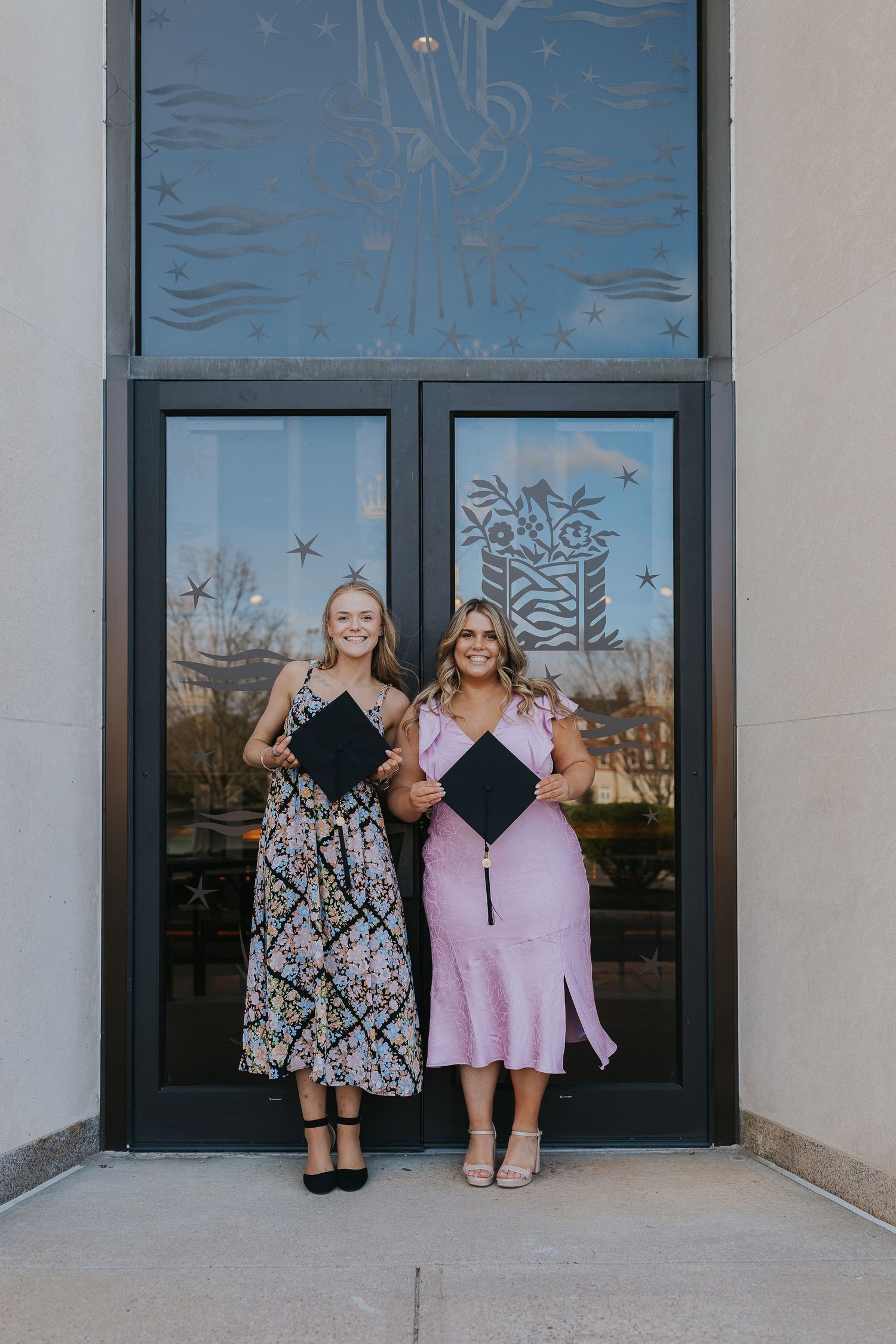 Two women in graduation attire holding caps outside university building.