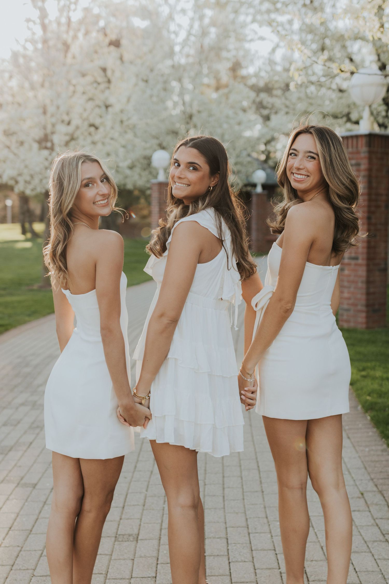 Three young women in white dresses holding hands and smiling near blooming cherry blossom trees.