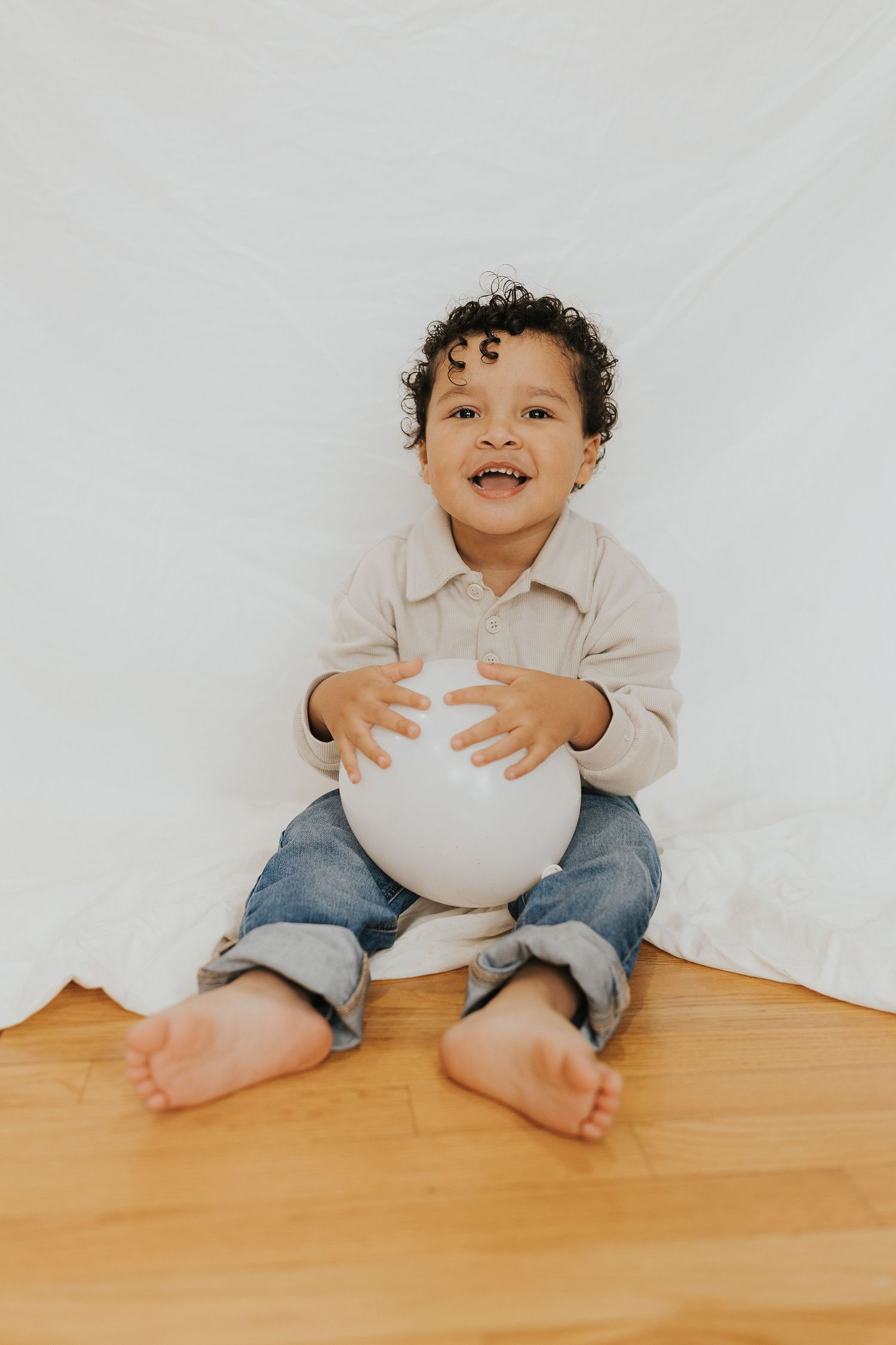 A happy barefoot toddler sits on a wood floor holding a white balloon against a white backdrop.