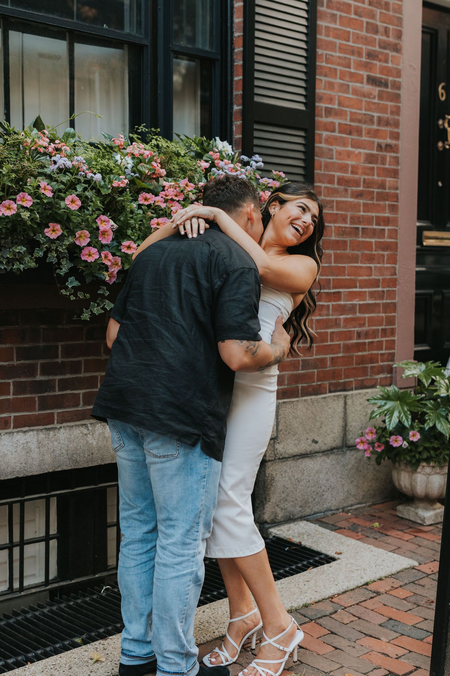 Joyful couple sharing a warm hug in front of a brick building adorned with colorful flower boxes.