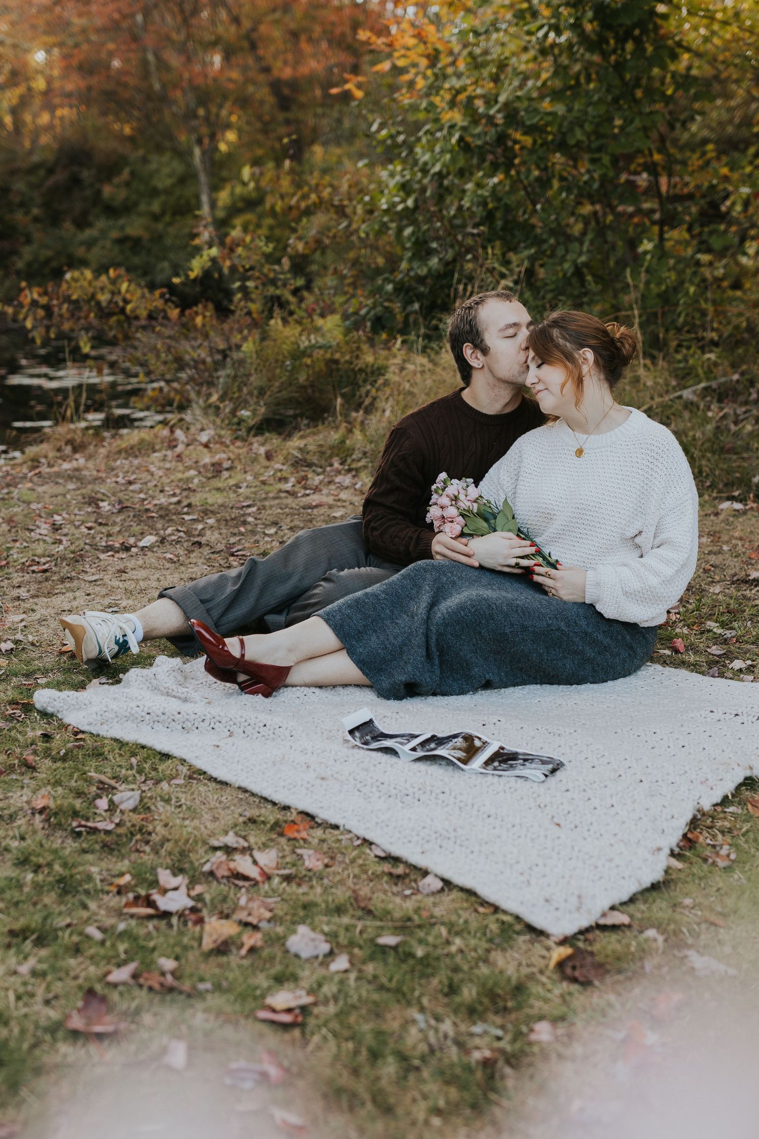 Couple sitting on a blanket in autumn park, sharing a kiss while holding flowers and ultrasound photos.