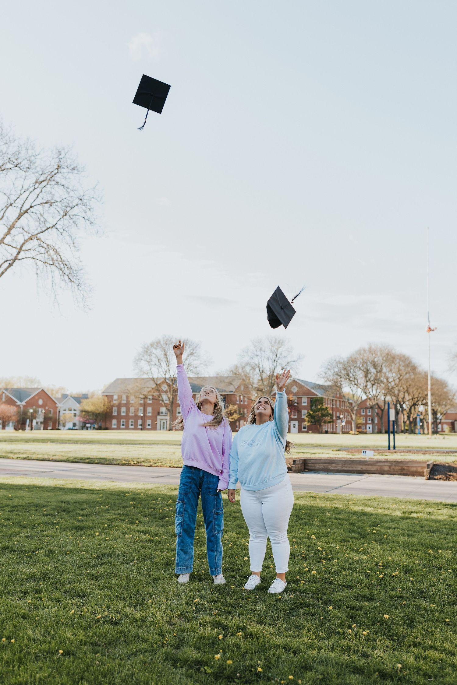 Two women tossing graduation caps in the air on green campus lawn celebrating achievement.