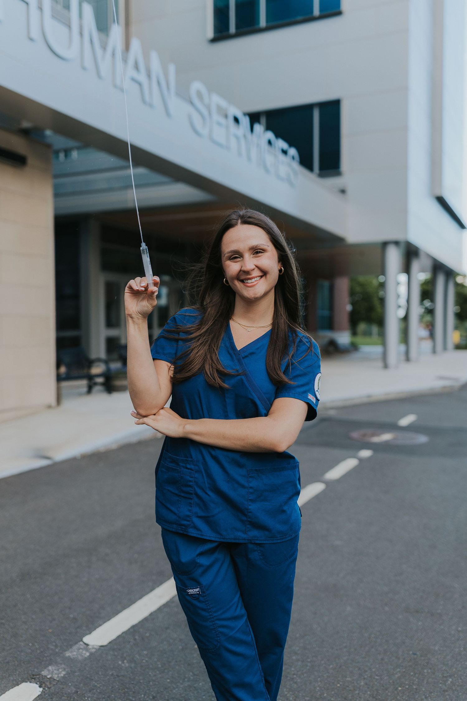 Smiling nurse in blue scrubs holding syringe outside Human Services building.