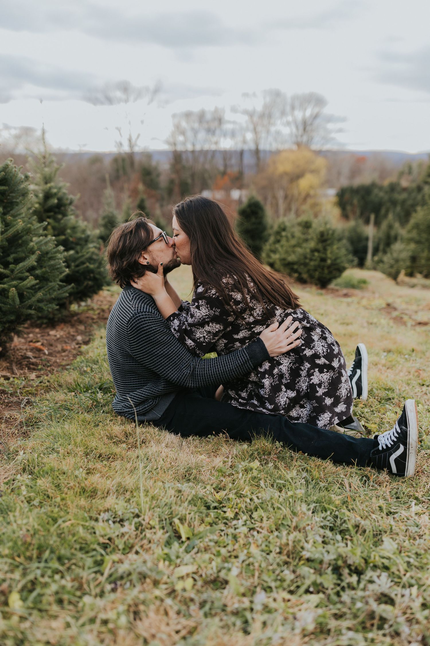 Couple shares a romantic kiss while sitting on a grassy hill at a Christmas tree farm.