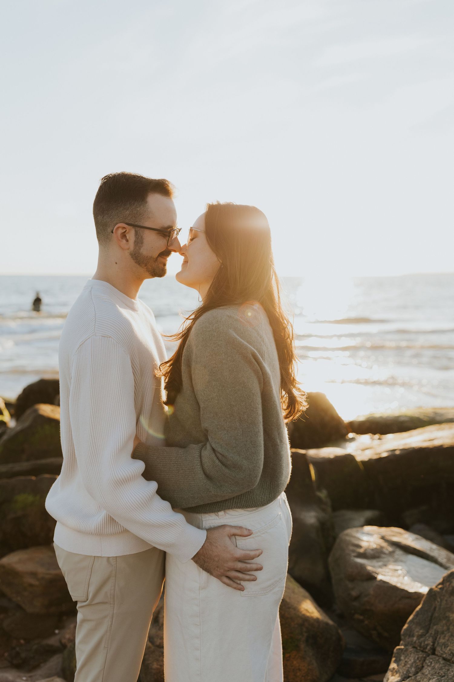 Couple sharing a romantic almost-kiss on rocky beach at golden sunset, man in white sweater embracing woman in gray top.