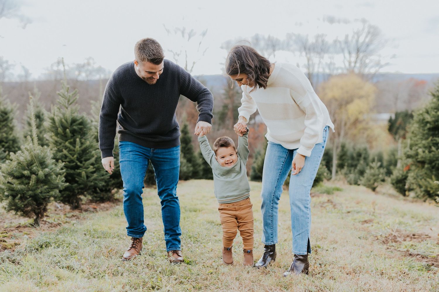 Two parents joyfully swing their toddler between them at a Christmas tree farm during a family session.