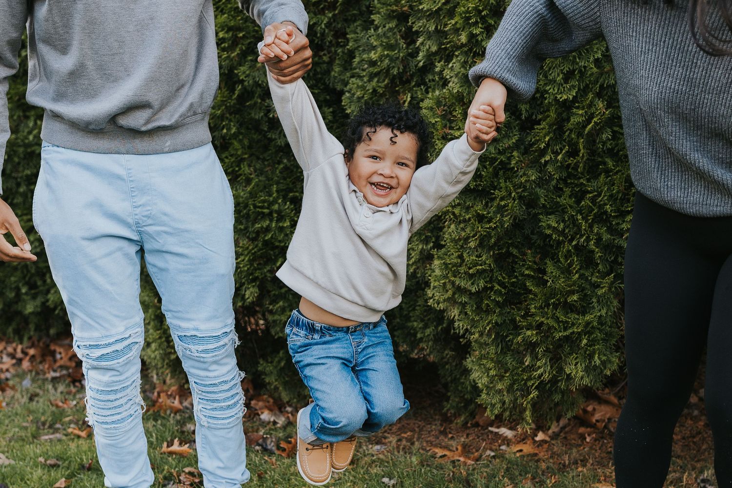 Joyful toddler being swung by two adults holding hands outdoors near green bushes on a sunny day.
