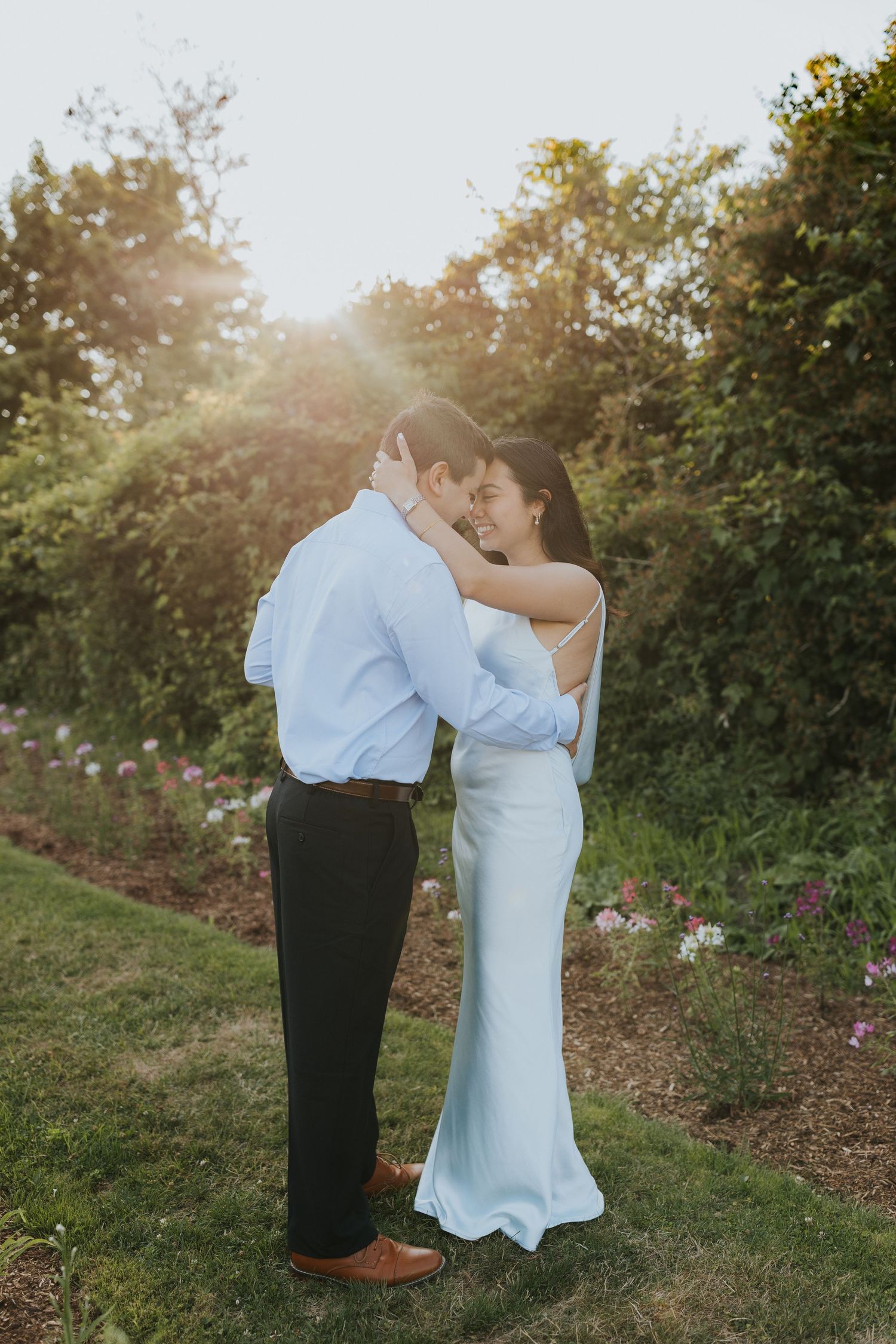 Couple embracing in a sunlit garden, woman in white slip dress and man in blue shirt, surrounded by lush greenery.