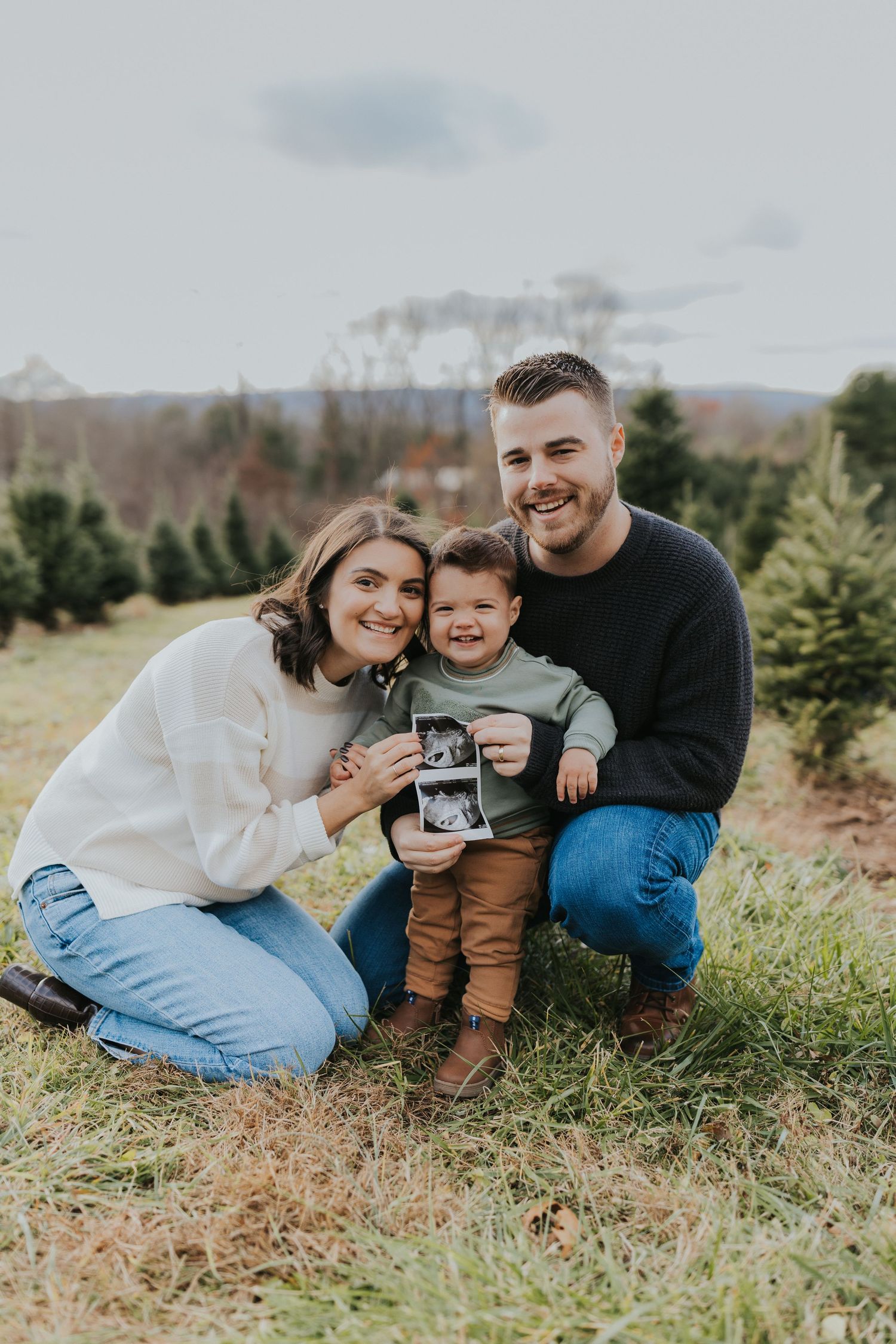 Smiling family posing at Christmas tree farm, young child holding ultrasound for pregnancy announcement.