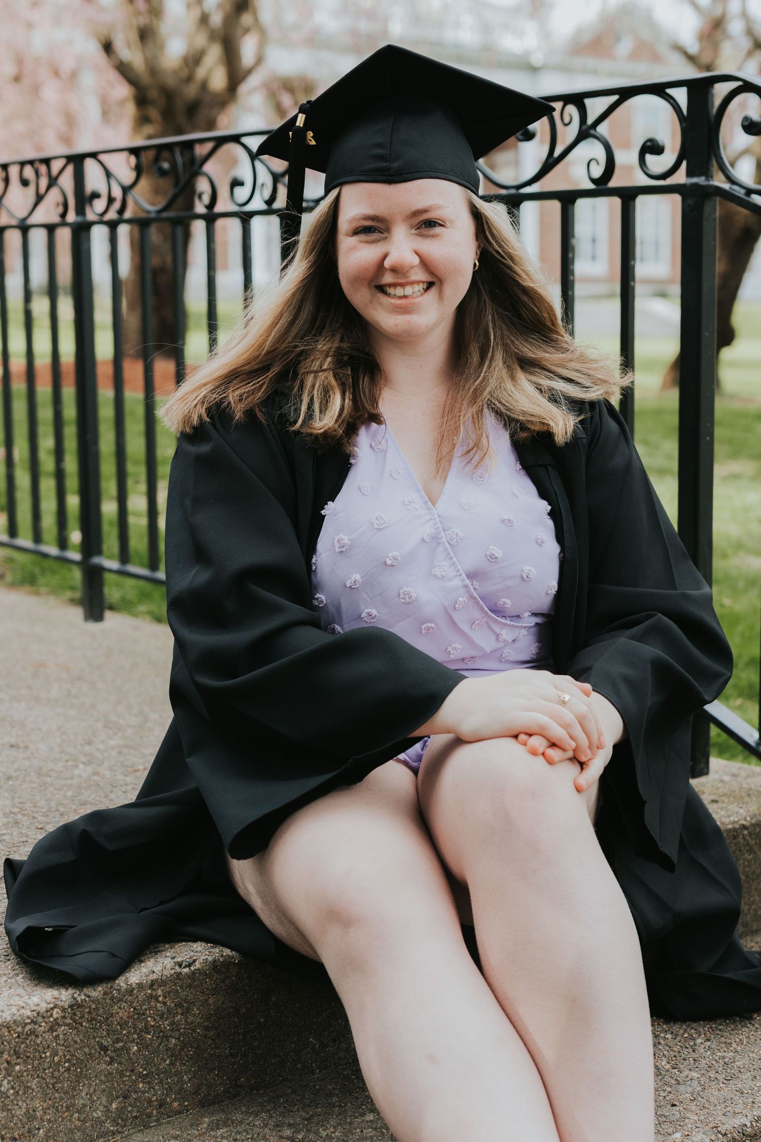 Cheerful graduate in black cap and gown sits on stone bench beside ornate iron fence on campus.