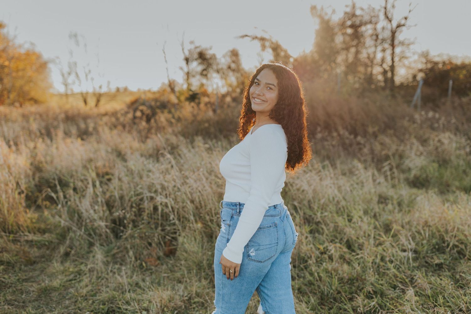Smiling young woman in white top and jeans posing in a golden autumn field at sunset.