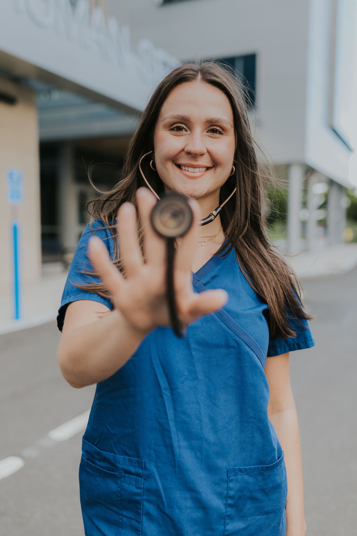 Smiling nurse in blue scrubs holding stethoscope toward camera outside medical building.
