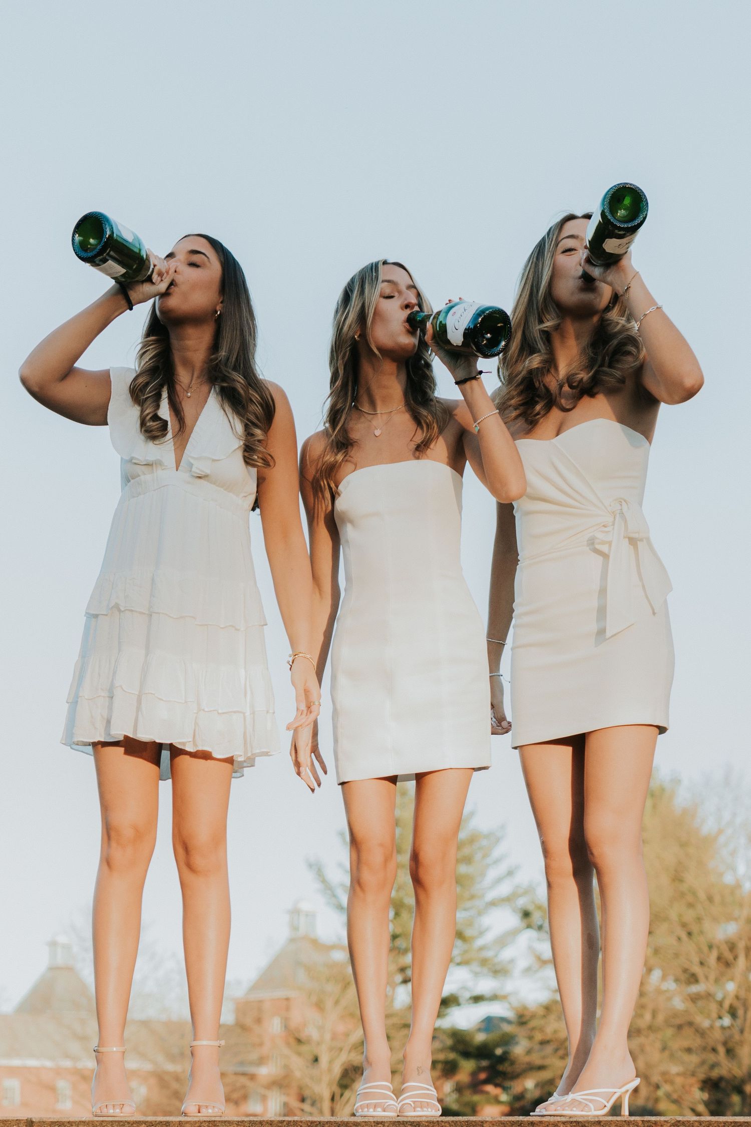 Three young women in white dresses celebrate graduation by drinking from champagne bottles outdoors on a sunny day.
