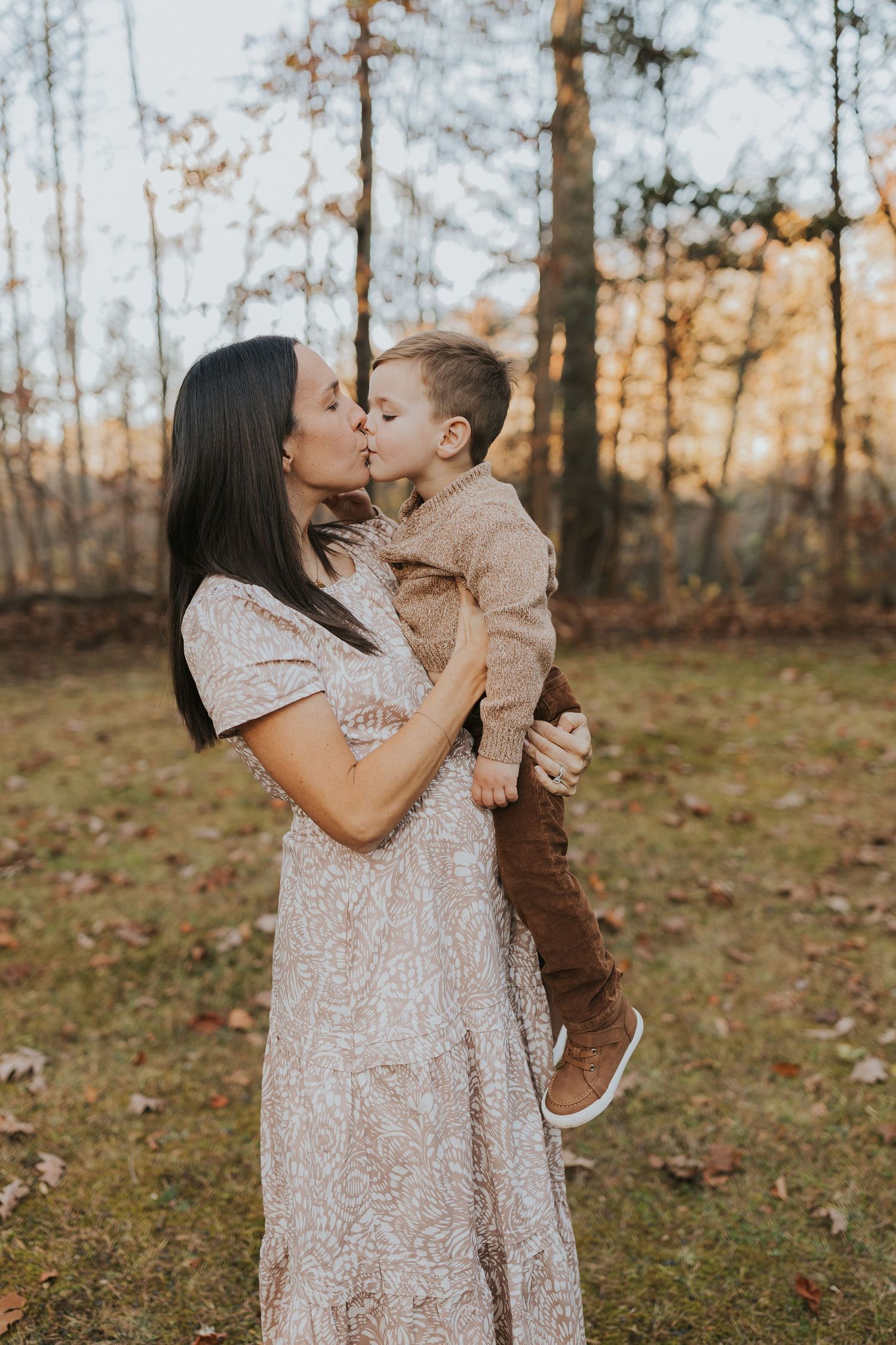 A dark-haired mother shares a tender kiss with her young son in a golden autumn forest.