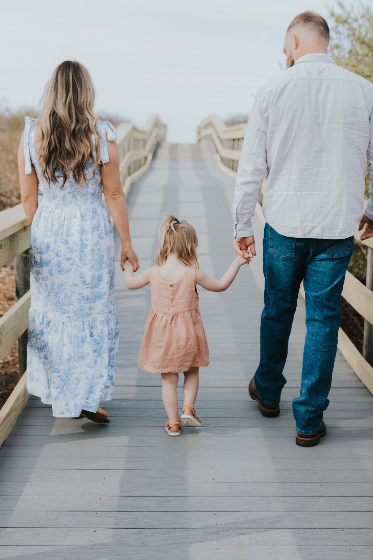 Family of three walking away on wooden boardwalk, parents holding young daughter's hands in matching outfits.