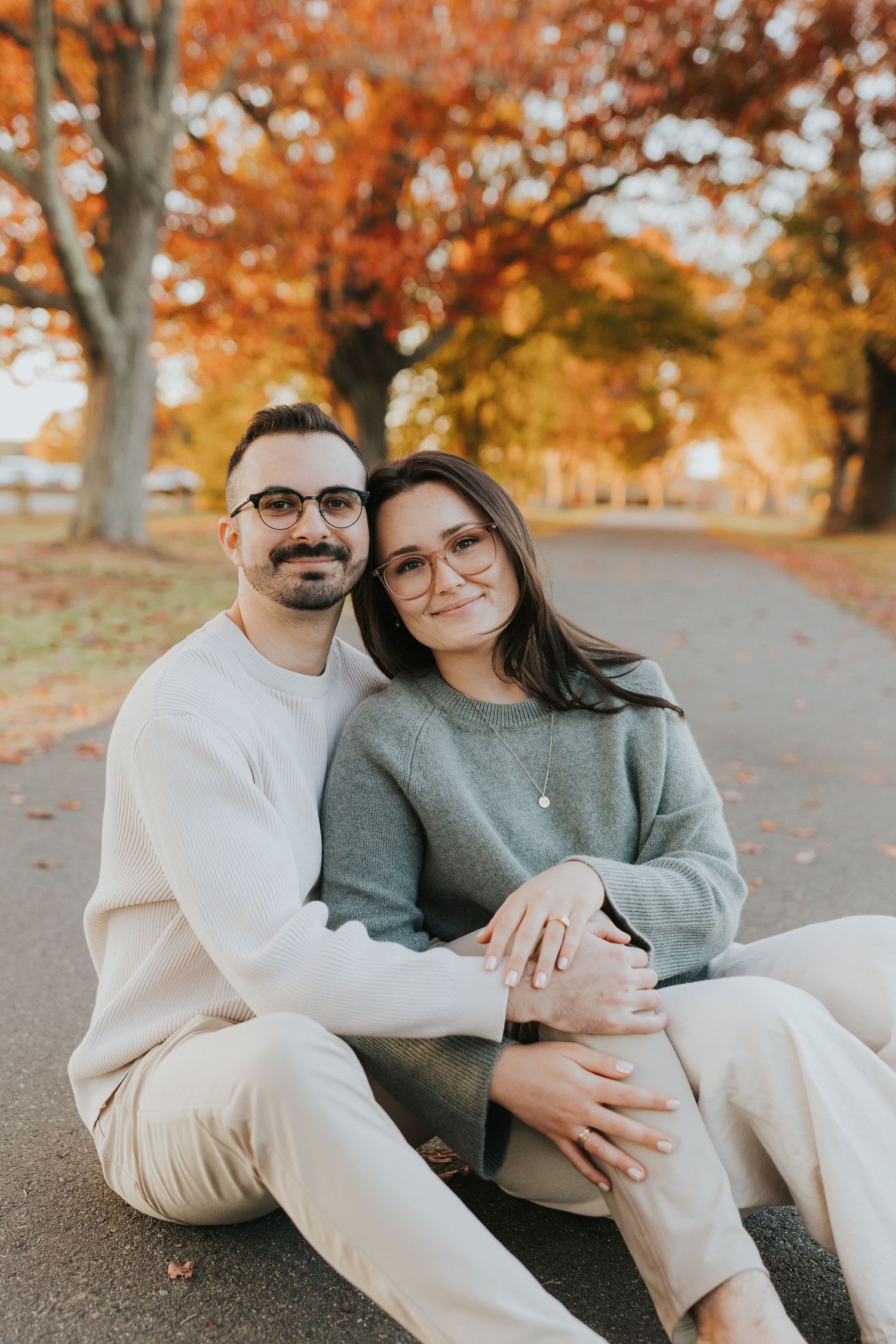 Couple sitting on autumn park path surrounded by colorful fall foliage, smiling at camera.