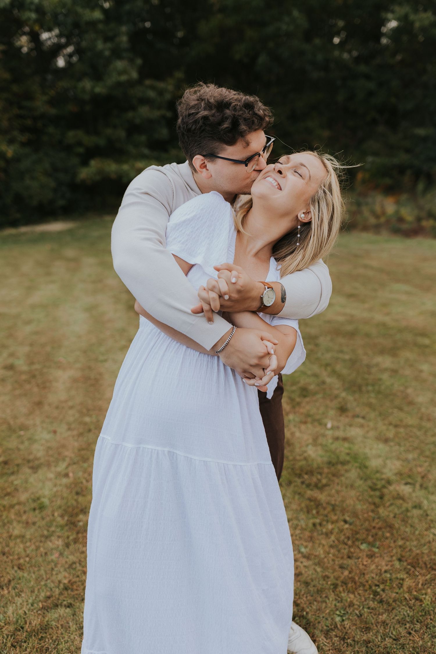 Man embracing and kissing woman in white dress during outdoor couples photo session in park.