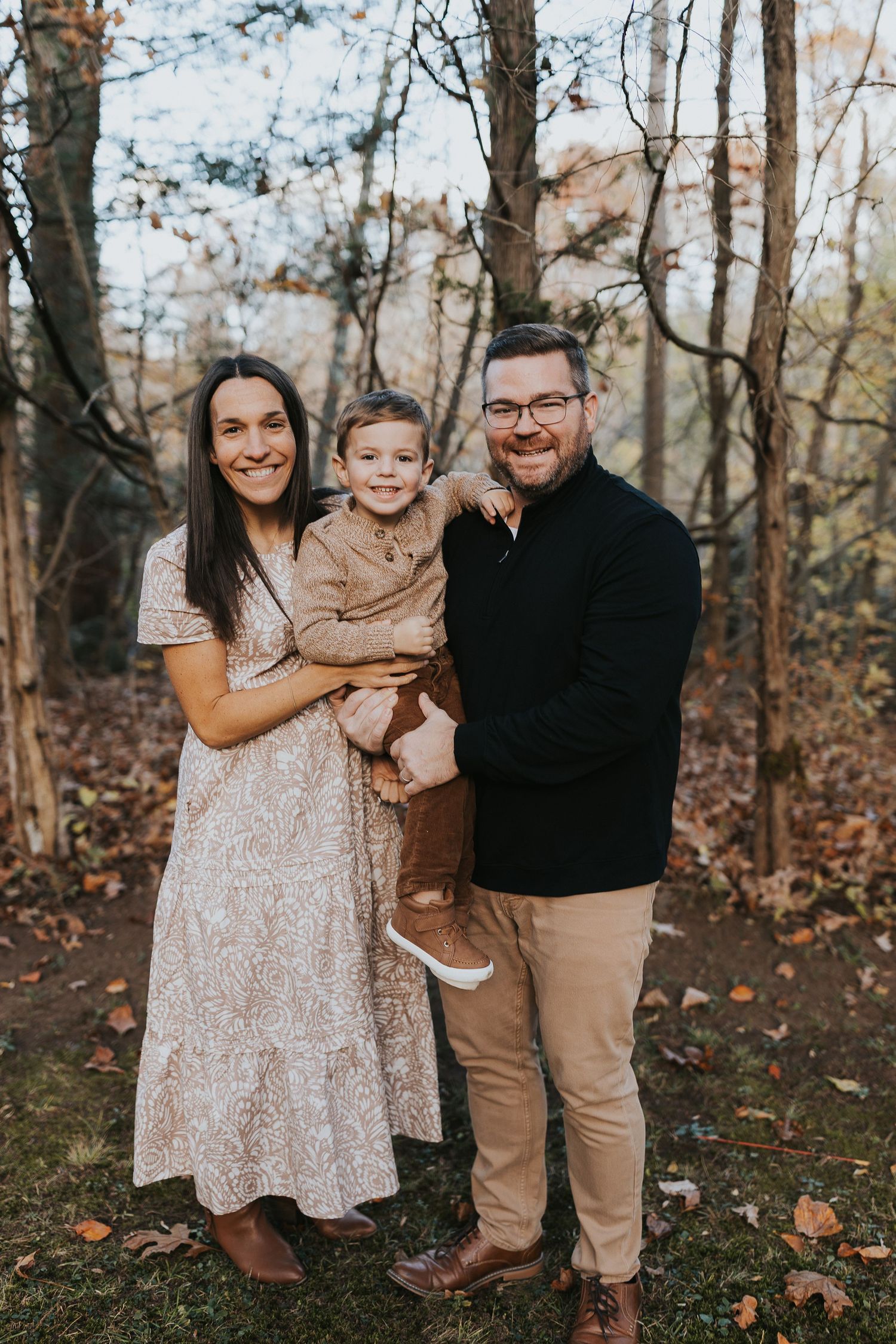 A happy family of three poses together in an autumn woodland setting, parents holding their young son.