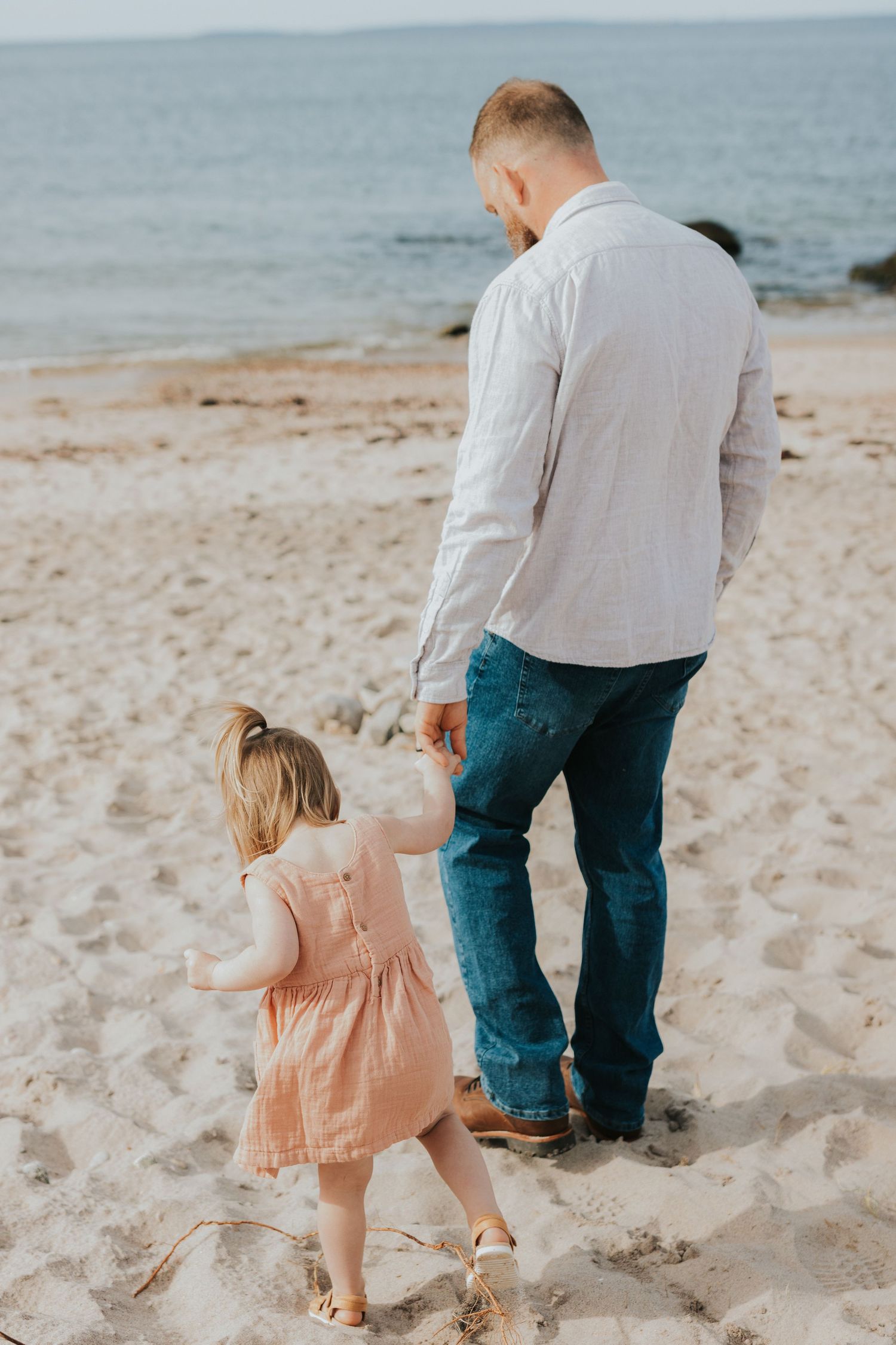 Bearded father in gray shirt walks hand-in-hand with toddler daughter on sandy beach.