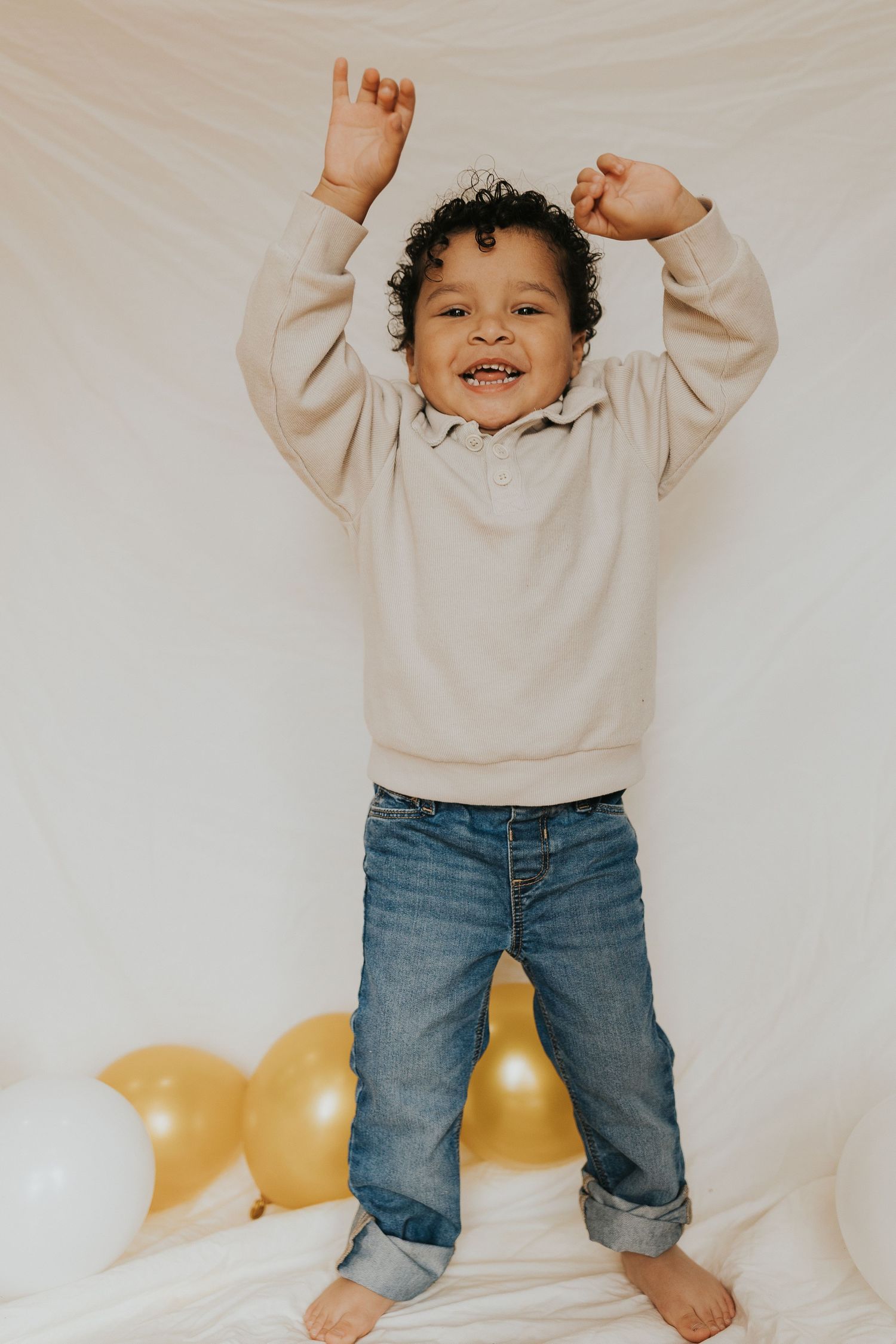 A joyful toddler in a beige sweatshirt and jeans celebrates with arms raised near gold balloons.