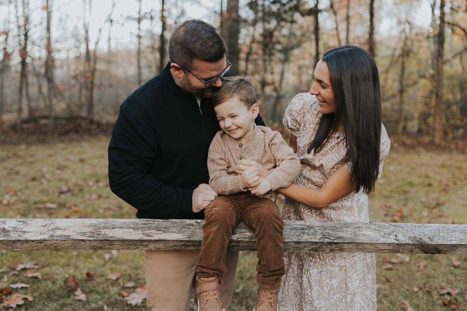 A happy family of three shares a tender moment together on a wooden fence in autumn woods.