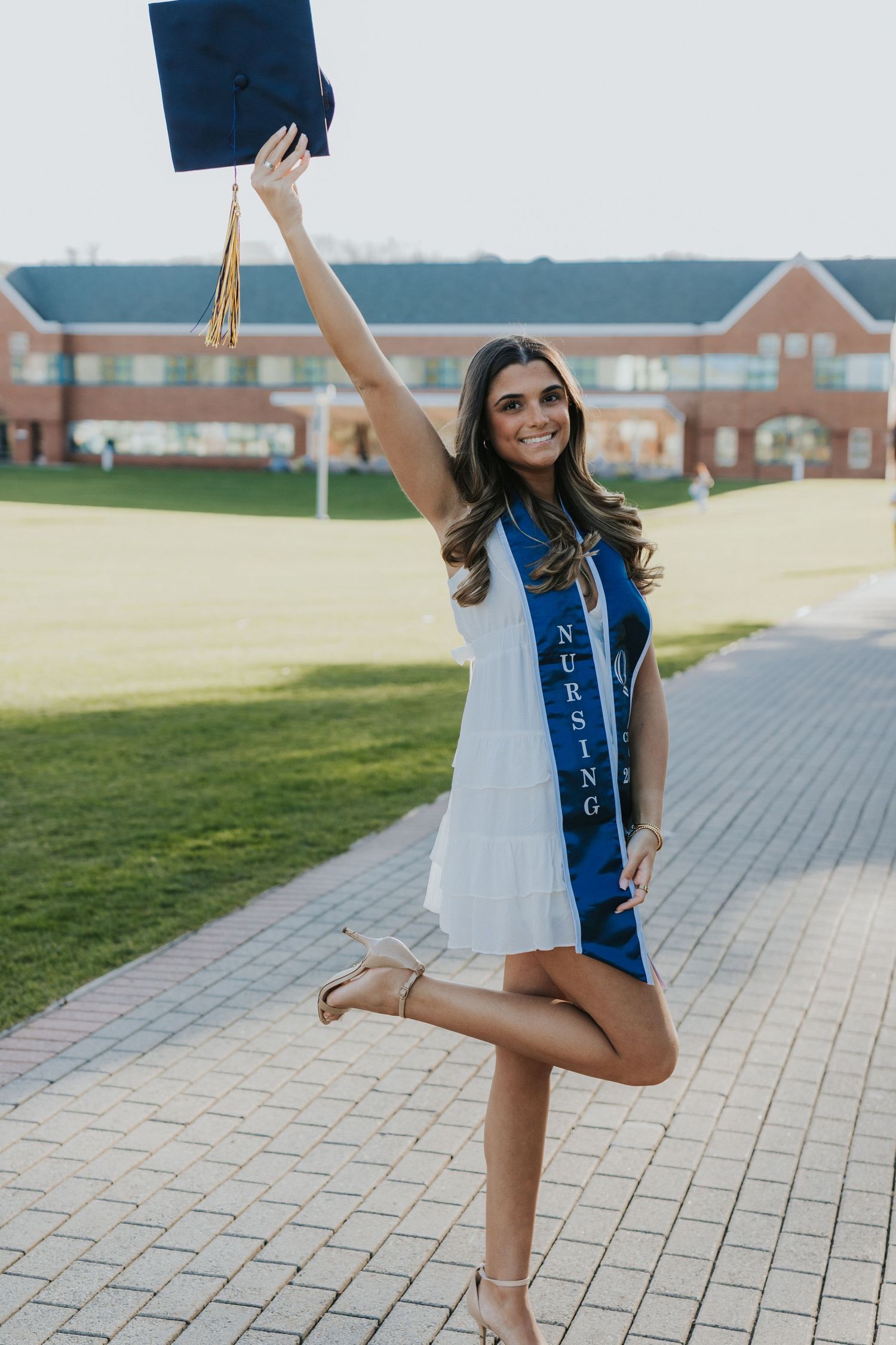 Nursing graduate in white dress celebrating with cap raised high wearing blue Nursing honor stole.