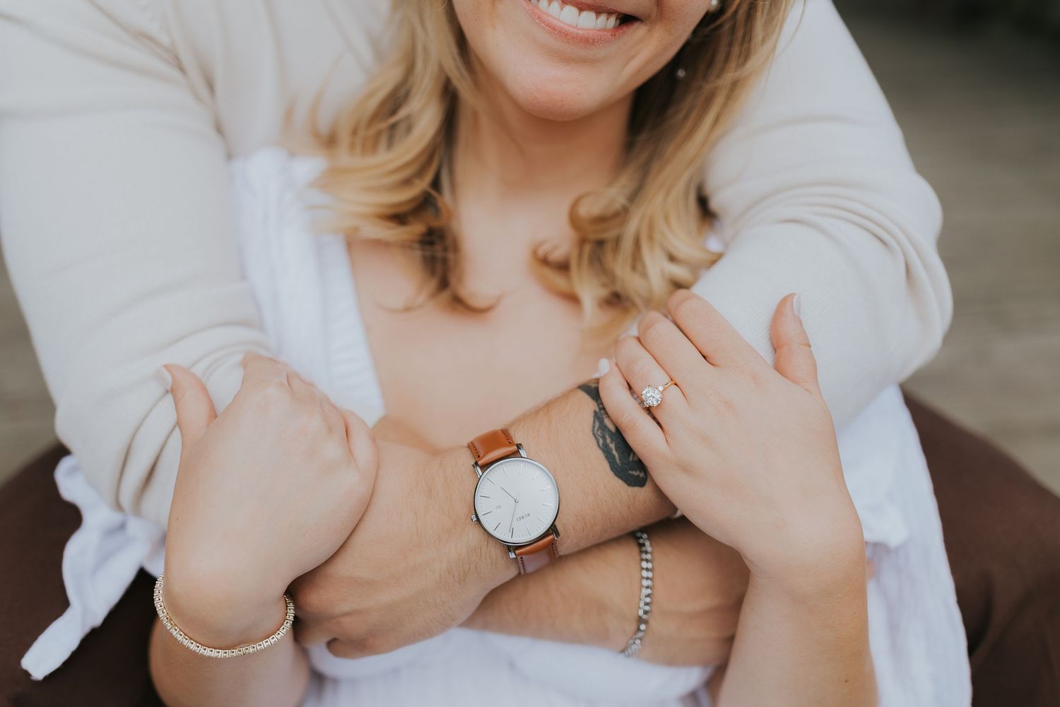 Close-up of smiling woman wearing a watch and ring, with arms wrapped around her in a tender embrace.