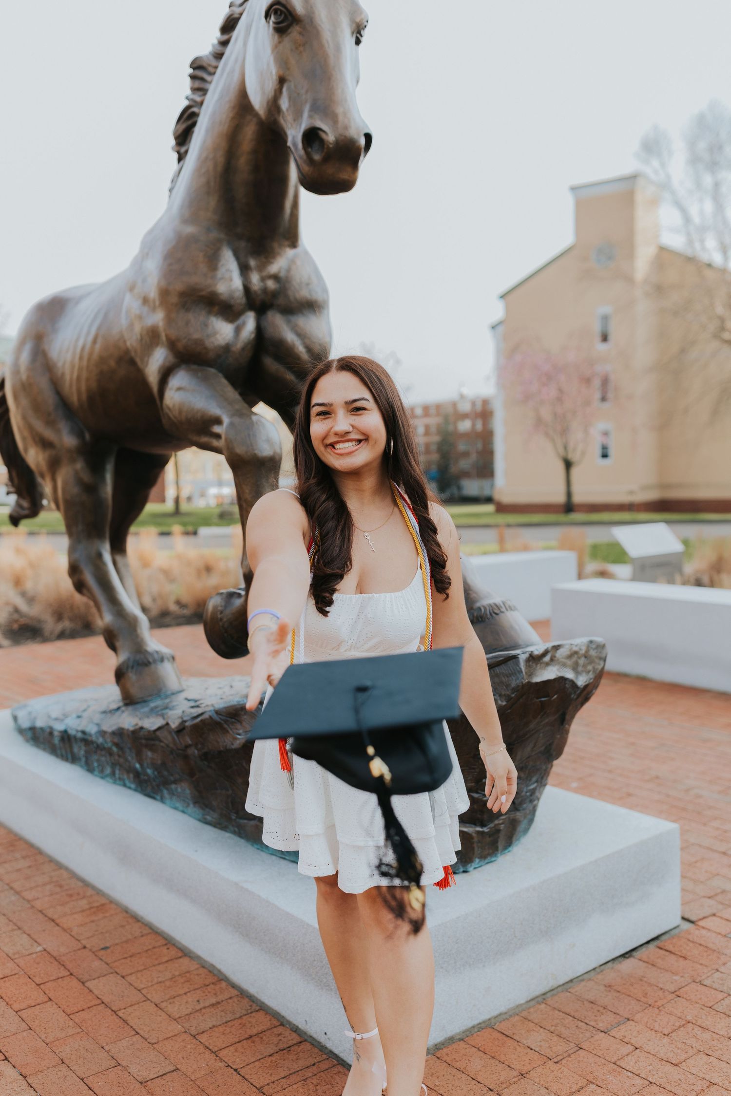 Smiling graduate in white dress holds cap in front of bronze horse statue on campus.