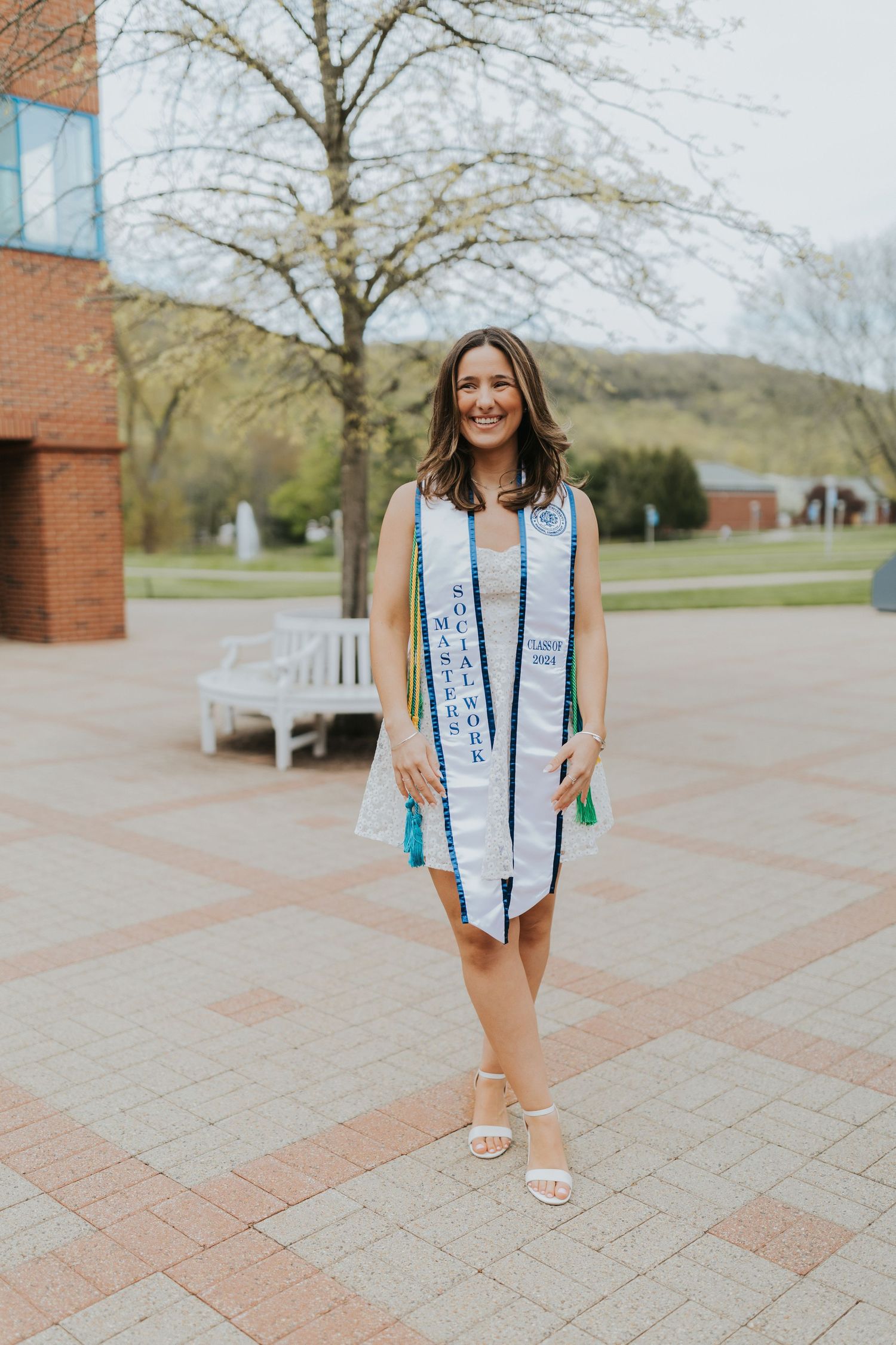 Smiling graduate stands on brick path wearing white stole and Master of Social Work sash outdoors.