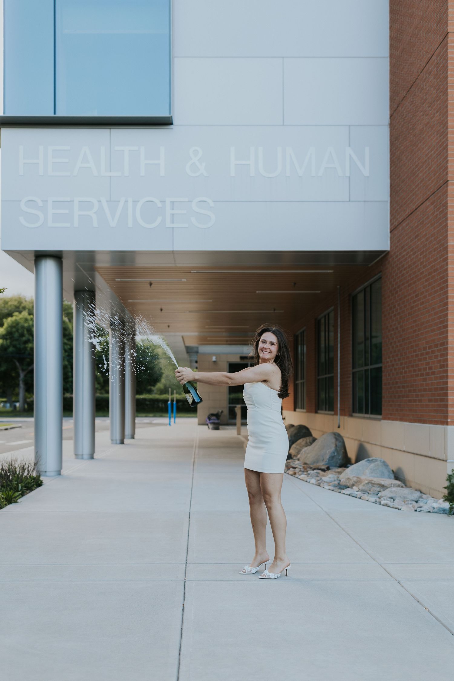 Nursing graduate pops champagne outside Health and Human Services building.