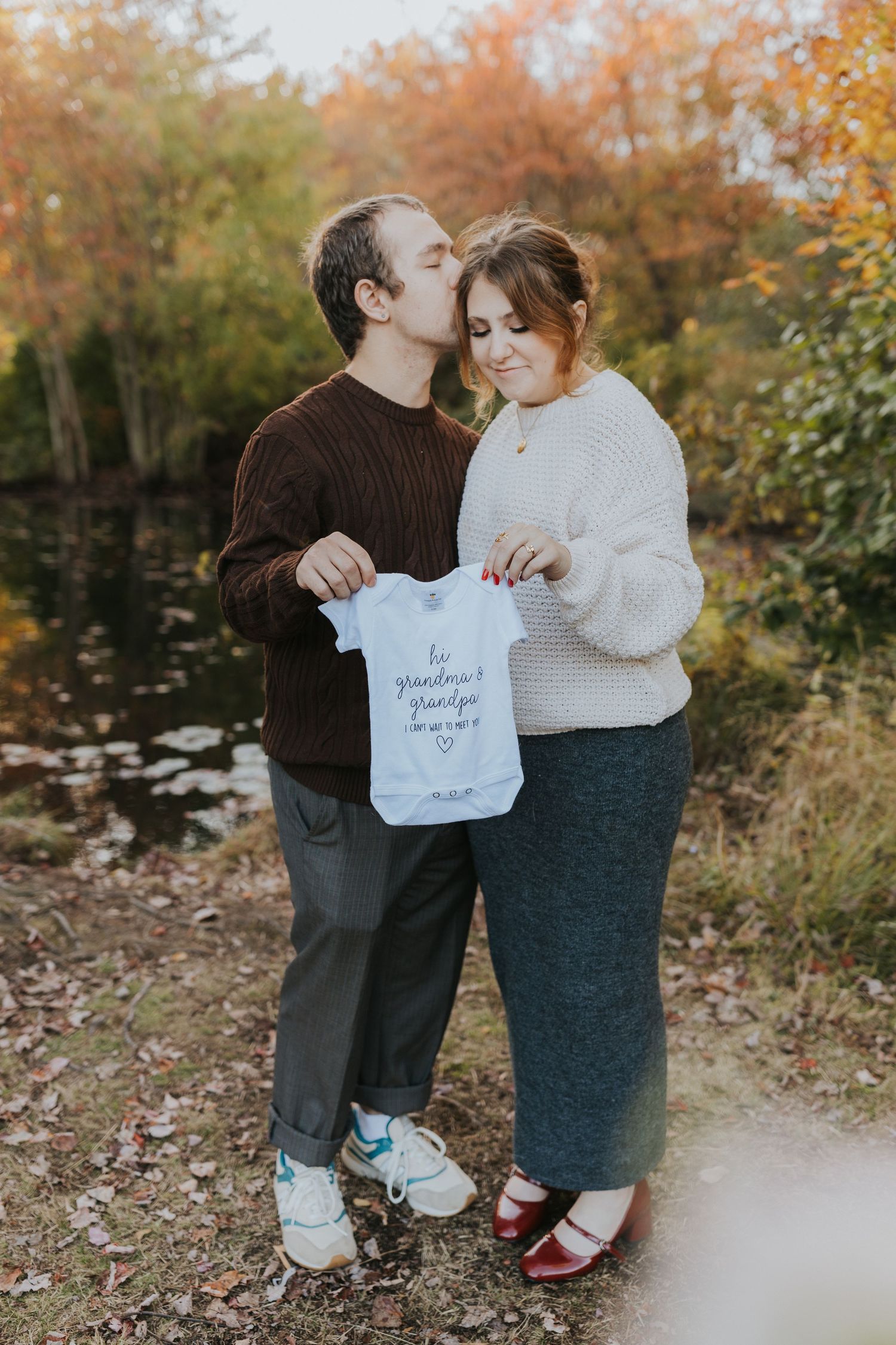 Couple kissing while holding baby onesie during outdoor fall pregnancy announcement photo session.