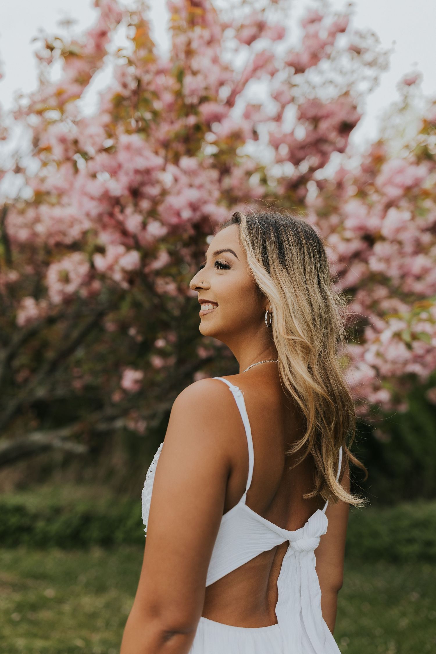 Young woman in white backless dress smiling near pink cherry blossom tree outdoors.