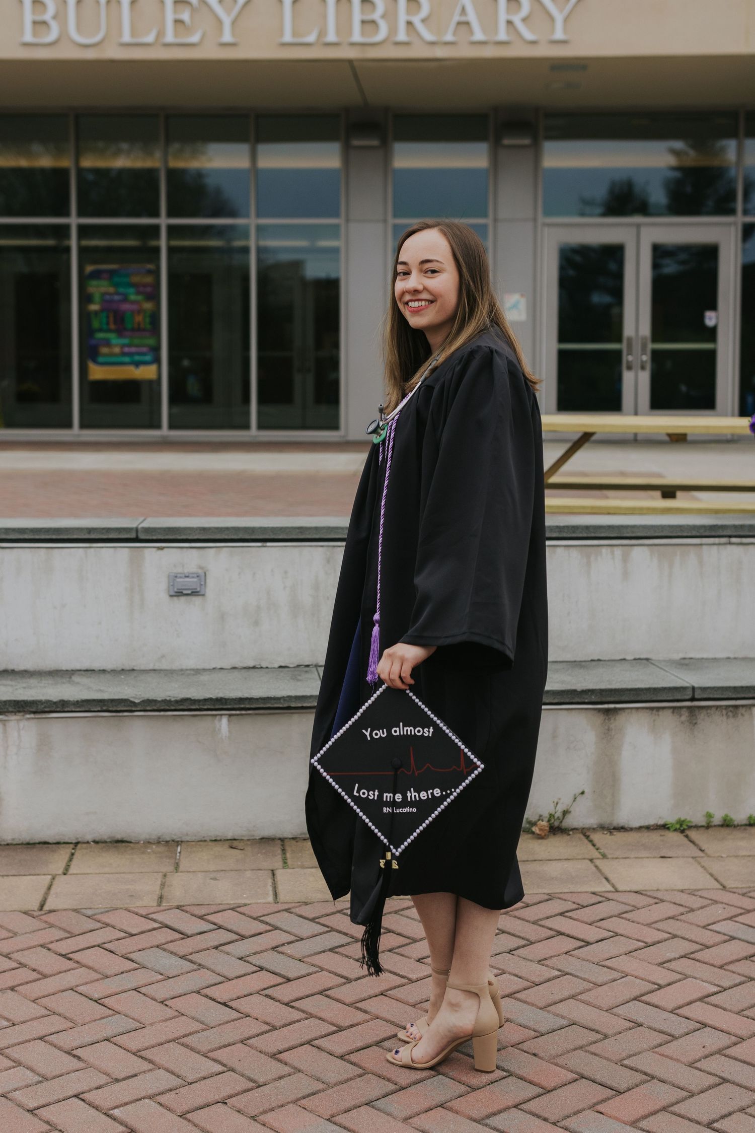 Female nursing graduate in black gown displays decorated cap reading you almost lost me there outside Buley Library.