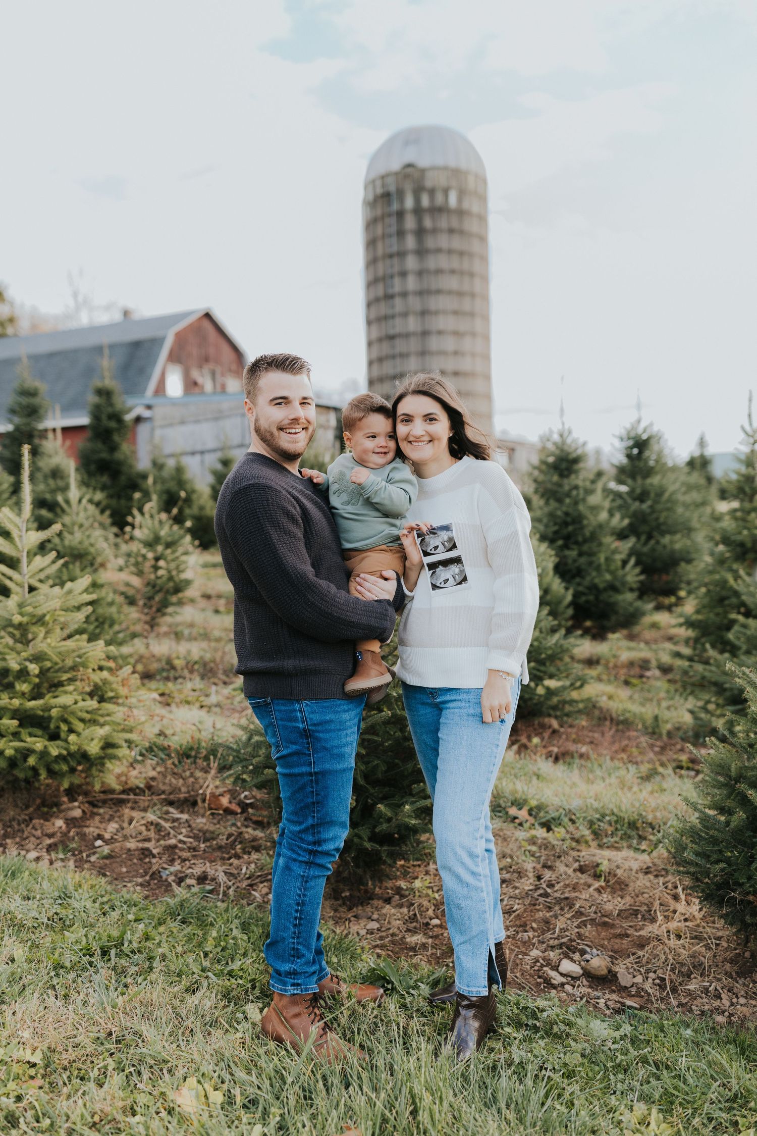 A young couple holds their toddler while displaying an ultrasound photo at a Christmas tree farm.