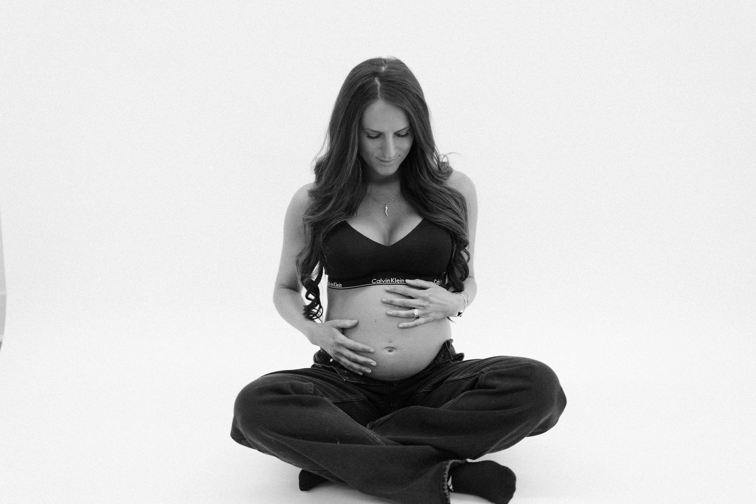 Black and white photo of a pregnant woman sitting cross-legged, cradling her baby bump with both hands.