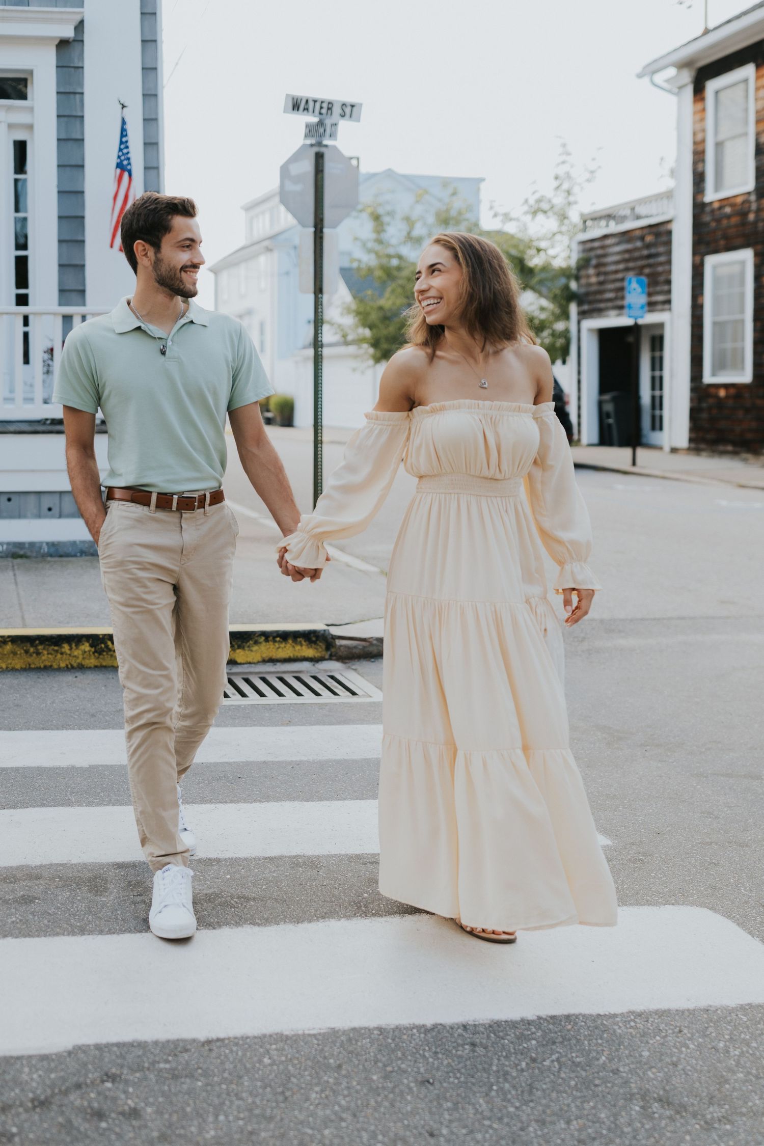 Smiling couple holds hands while crossing a street in a charming coastal town, dressed in summer outfits.