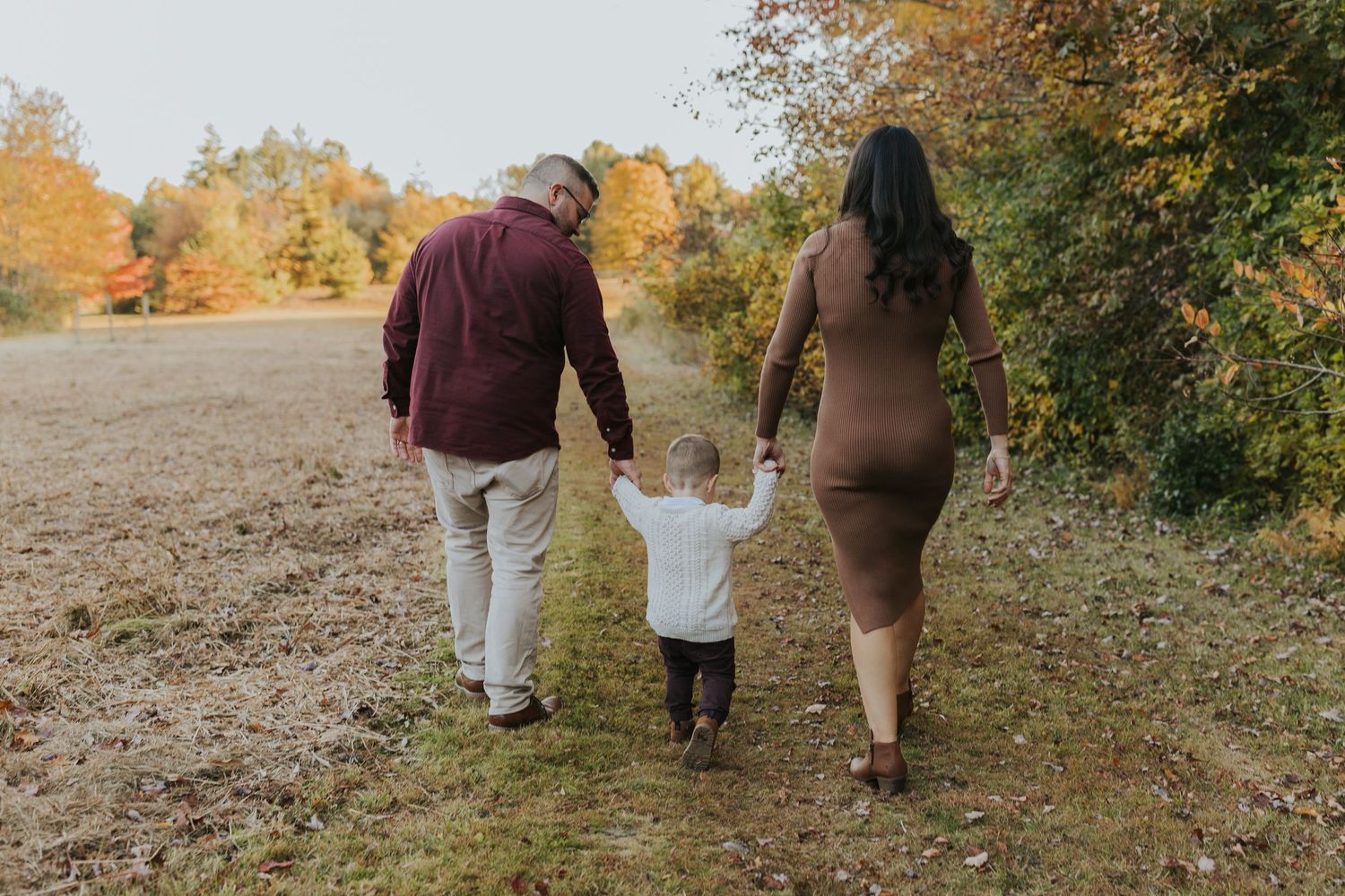 Young family walks hand-in-hand through autumn field, parents guiding toddler between them.